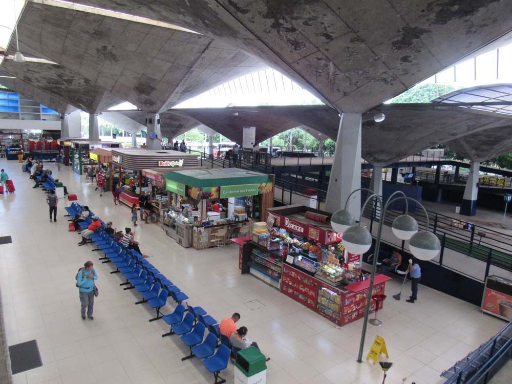 Interior de um terminal de transportes com barracas de comida, assentos e pessoas. Teto de concreto com formas triangulares.