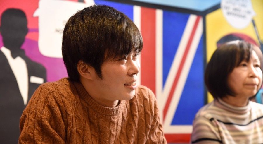 Two people seated indoors, one in a brown sweater and one in a striped turtleneck, with a colorful mural behind them.