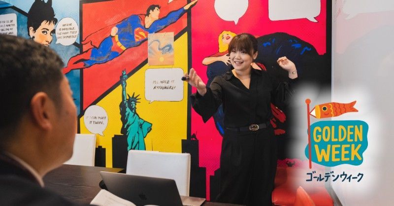 Presenter posing in front of a colorful “Golden Week” event display with superhero-themed graphics