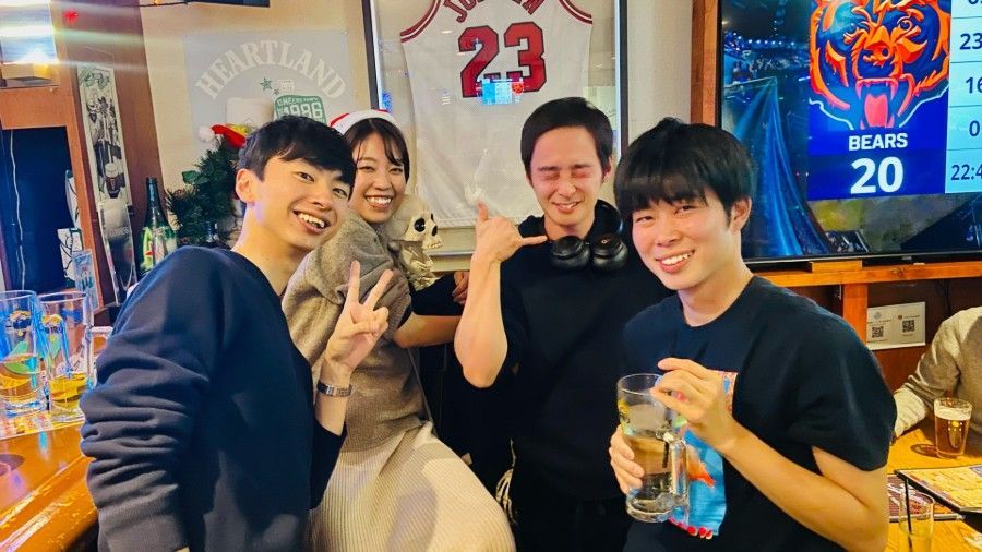 Four friends smiling at a restaurant table, holding drinks in a lively sports bar setting.