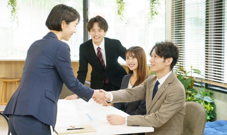 Two businesspeople shaking hands in a bright office while two colleagues smile nearby.