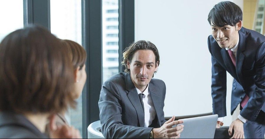 Business meeting with three people around a laptop in a bright office, one man speaking and another leaning in.