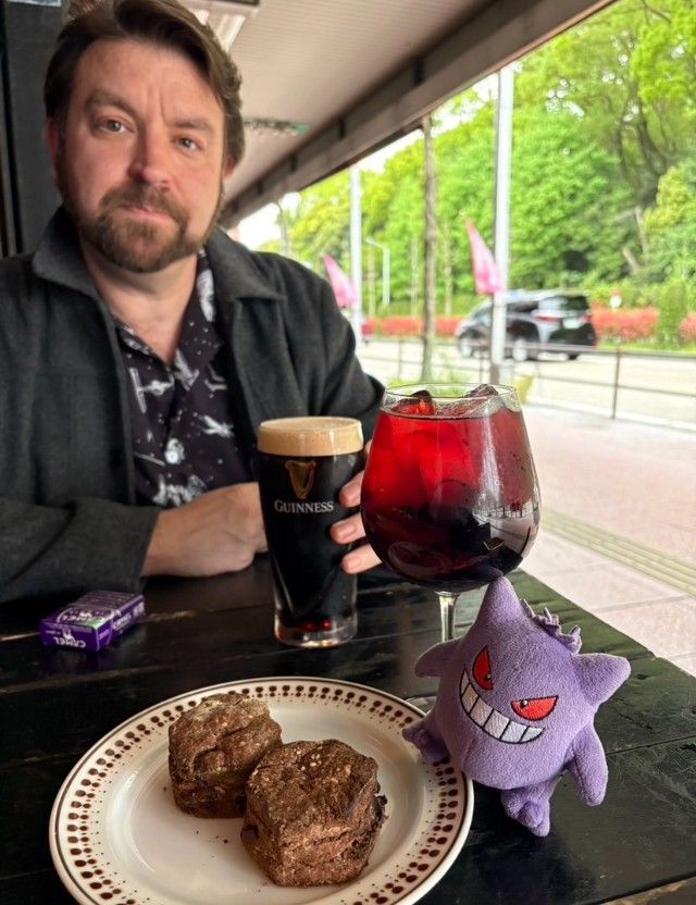カフェのテーブルに座る男性。テーブルにはペストリー、コーヒー、赤い飲み物、そして紫色のゲンガーのぬいぐるみが置かれている。
