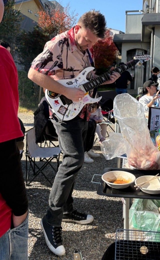 屋外の小さな集まりで、白いエレキギターを演奏している人がおり、近くではフライパンで料理をしている。