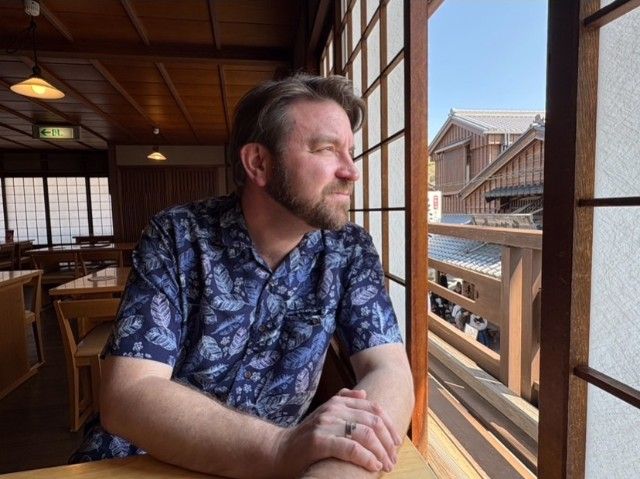 青い柄のシャツを着た男性が、木造のカフェのバルコニーから窓の外を眺めている。