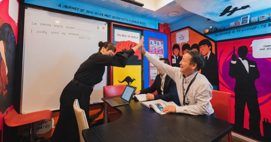 Two people high-five in a colorful classroom or office with a whiteboard and laptop, while others watch nearby