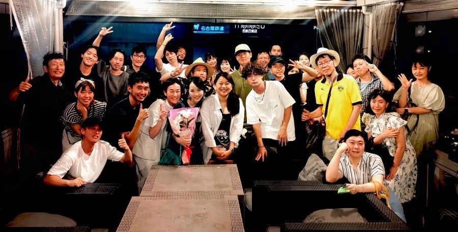 Large group posing together at a table in a festive indoor setting, smiling and waving at the camera.