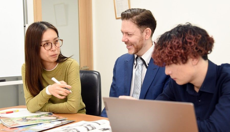 Three coworkers discuss a project at a table with a laptop and papers.