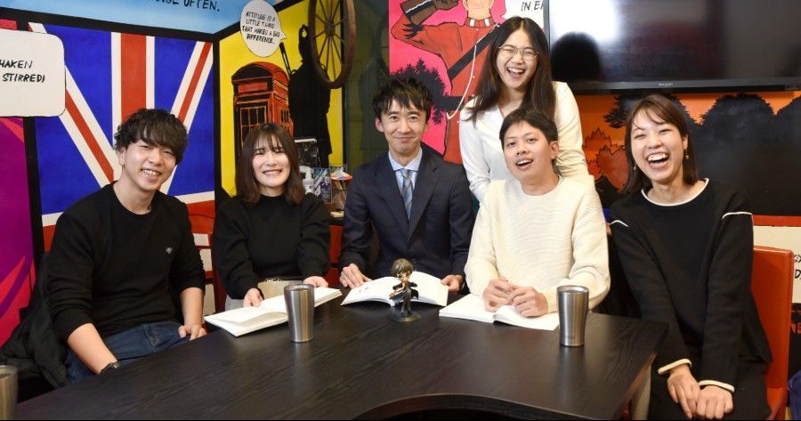 Six people seated at a table, laughing. The background features a Union Jack flag and colorful decor.