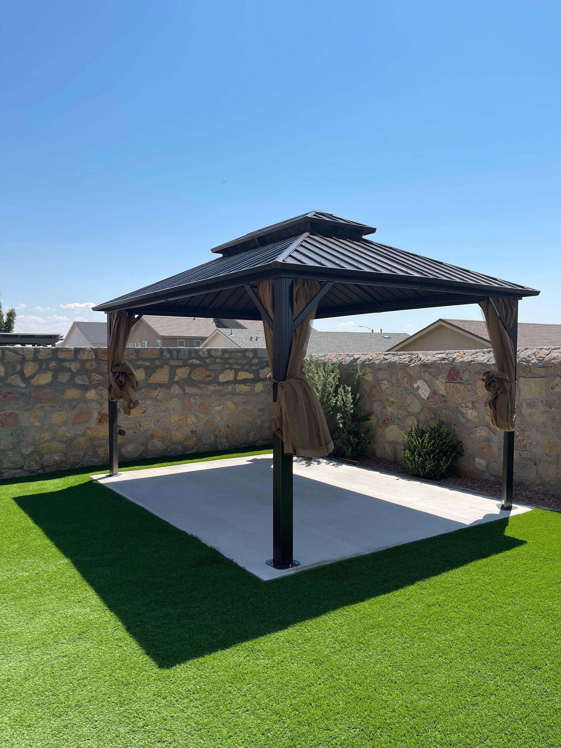Wooden gazebo with a dark roof on a white patio, surrounded by green grass and a stone wall.