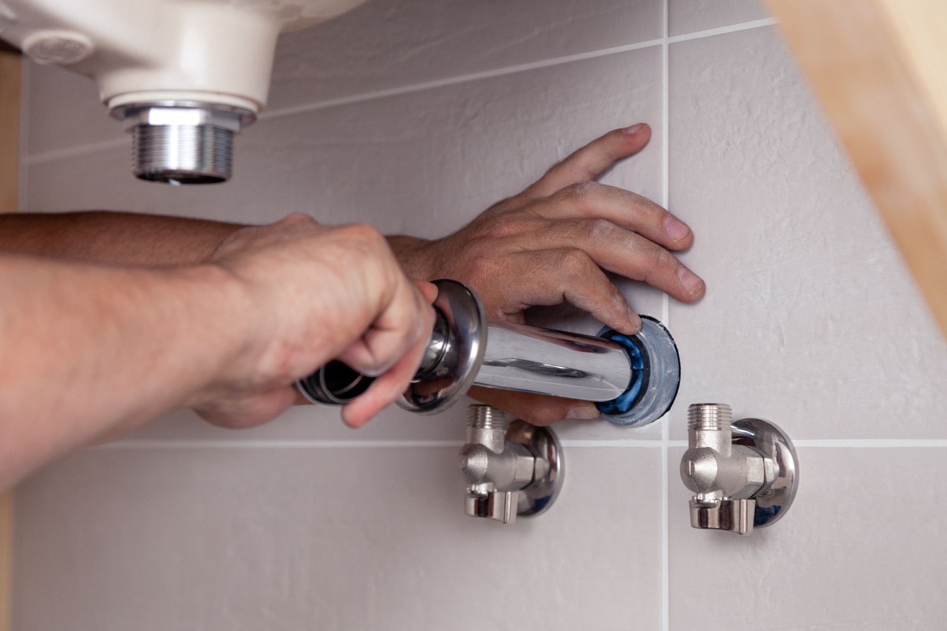 Hands tightening a chrome pipe fitting on a tiled bathroom wall under a sink.