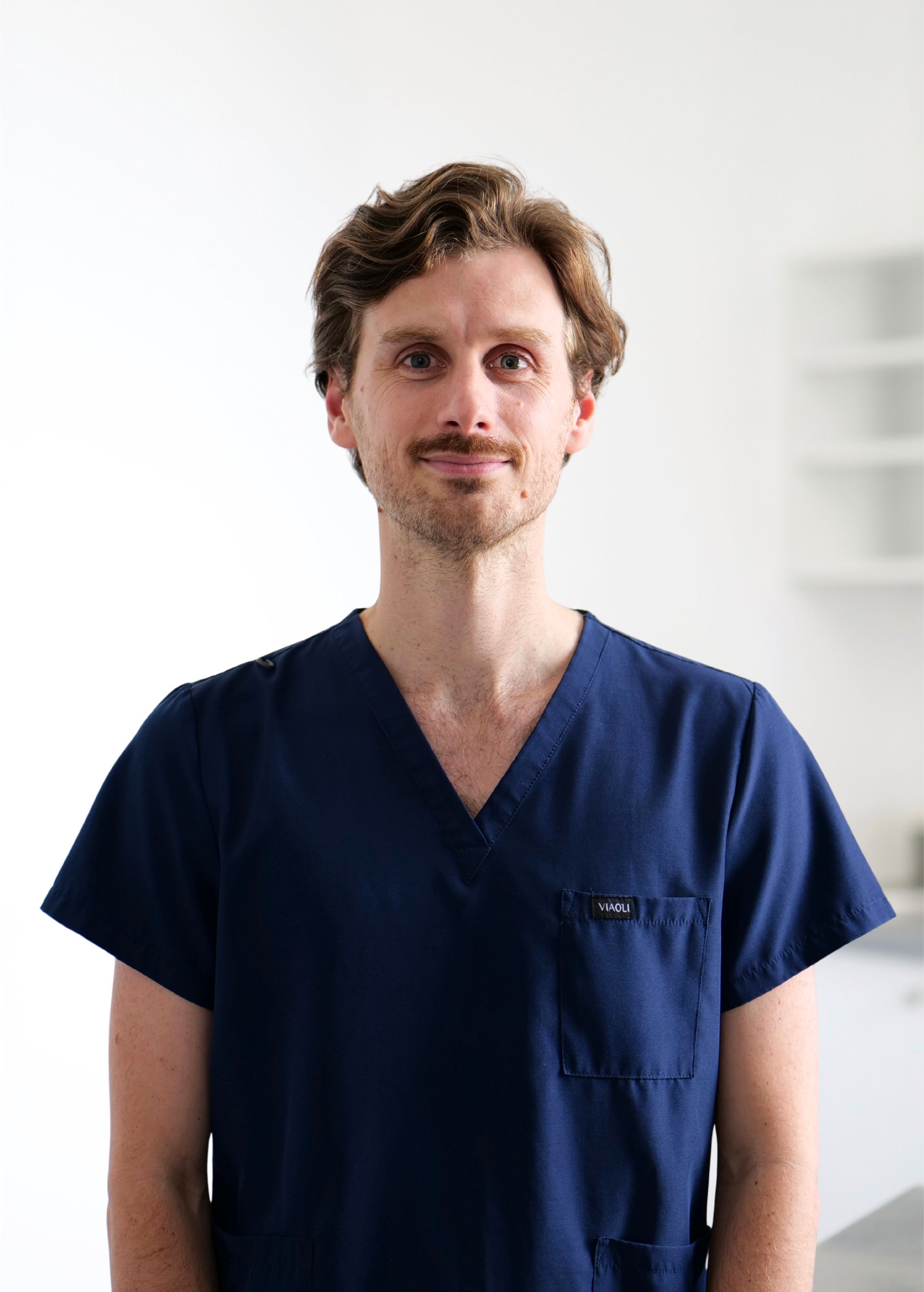 Personne en blouse médicale bleu marine, debout sur fond blanc, face à la caméra.