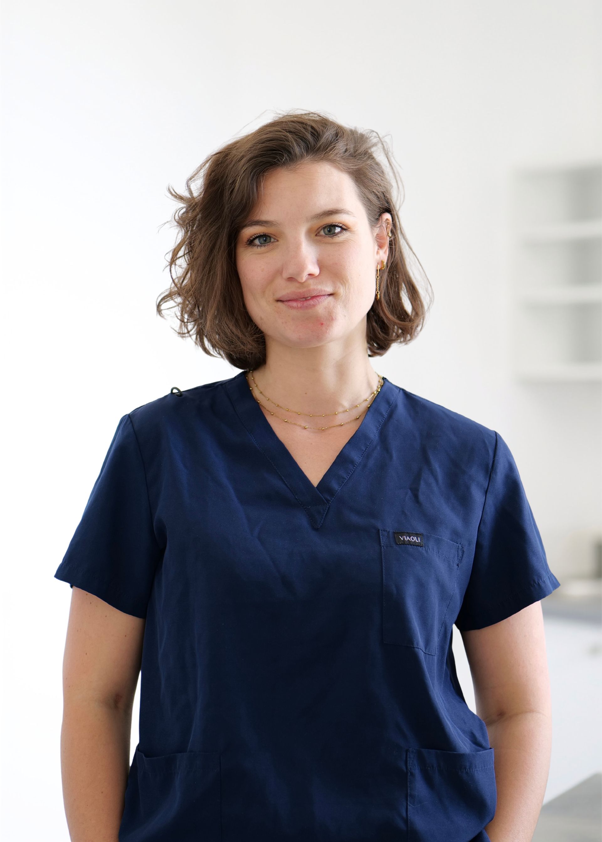 Femme en blouse médicale bleu marine, debout à l'intérieur sur fond clair, regardant droit devant elle avec une expression neutre.