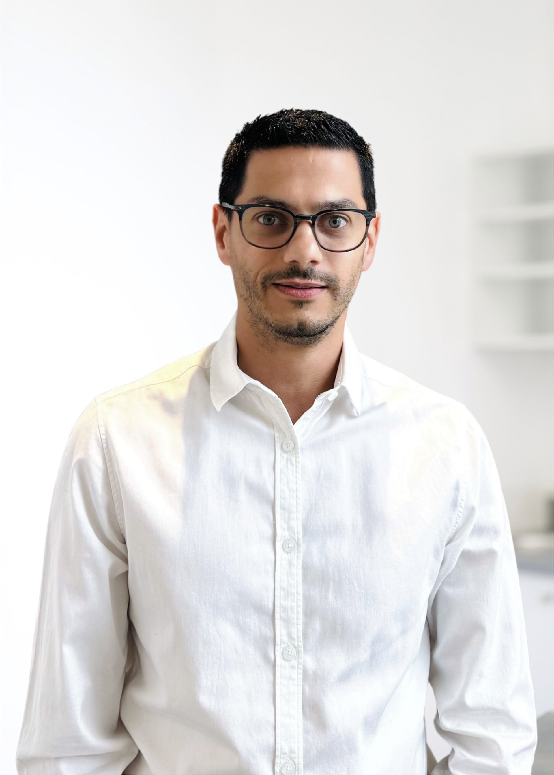 Homme debout à l'intérieur, portant une chemise blanche boutonnée et des lunettes, sur un fond uni et lumineux.