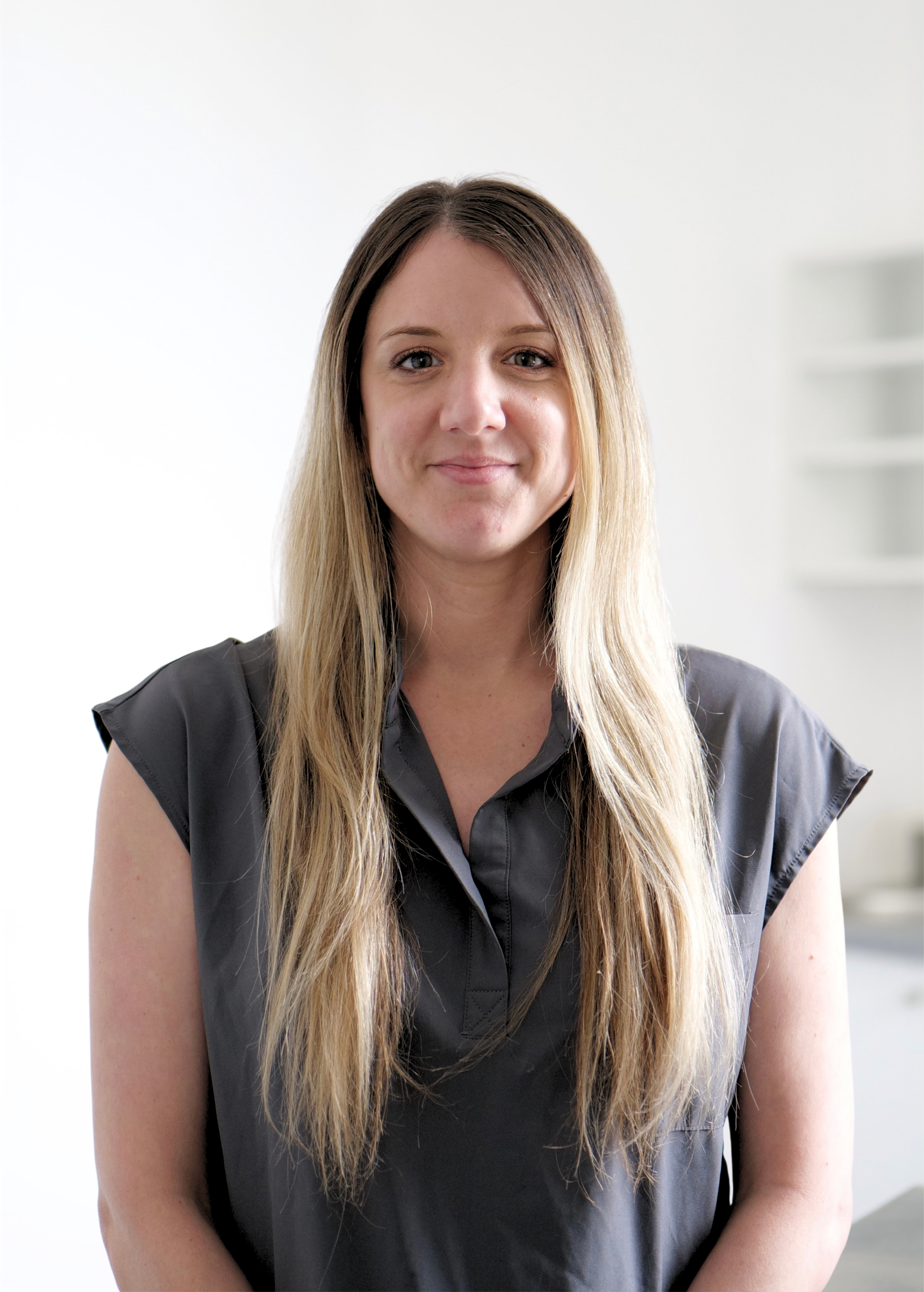 Portrait d'une femme aux longs cheveux blonds, vêtue d'un chemisier sombre sans manches, se détachant sur un fond blanc éclatant.