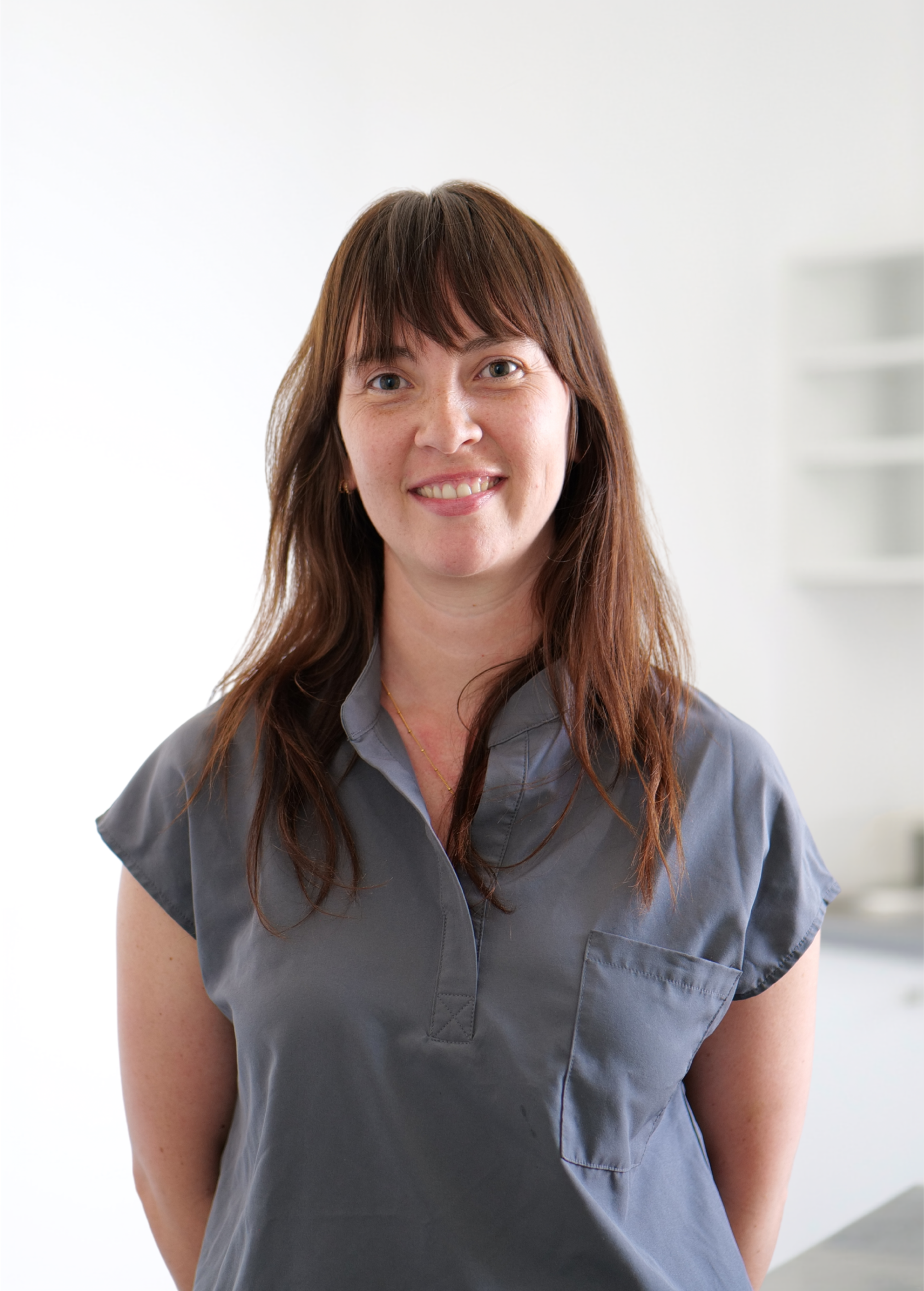 Femme debout à l'intérieur, sur fond blanc, vêtue d'un chemisier gris et souriant à l'objectif.