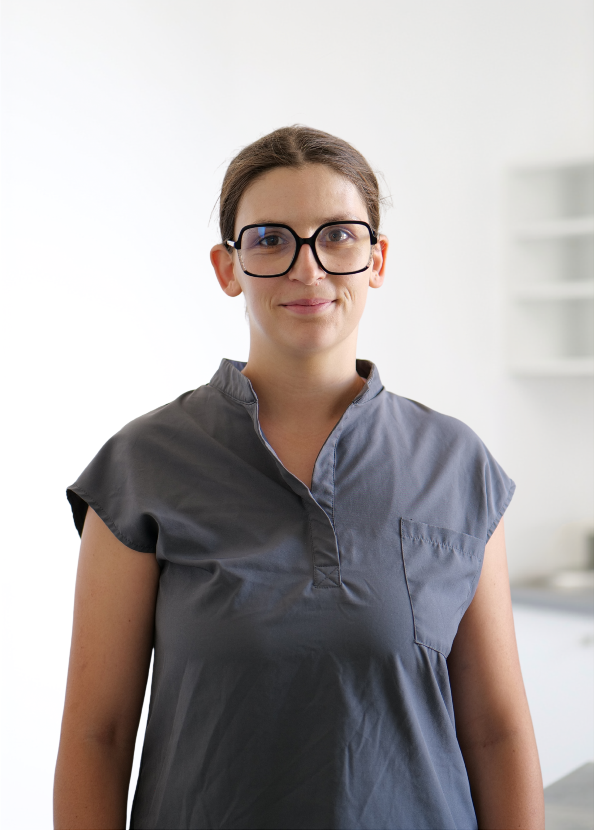 Personne portant des lunettes et une chemise grise, debout à l'intérieur sur un fond lumineux.
