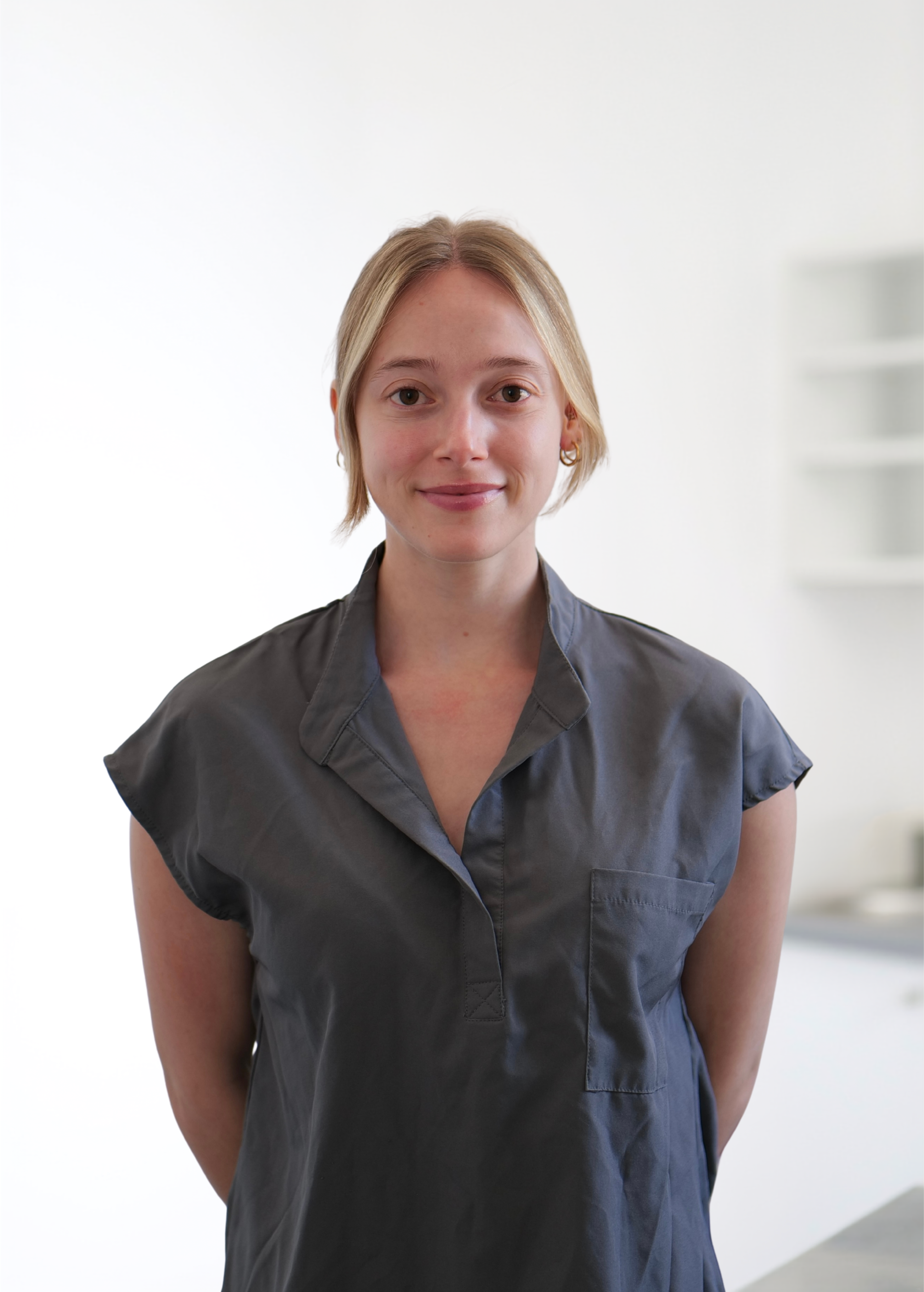 Une personne se tient debout à l'intérieur, vêtue d'une chemise gris foncé et souriant dans une pièce lumineuse.