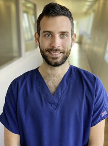 Une personne souriante en blouse bleue se tient dans un couloir d'hôpital.
