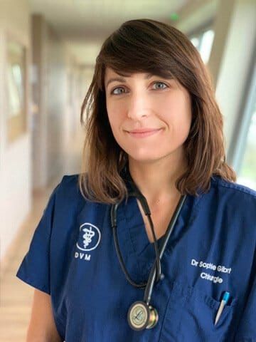 Une soignante en blouse bleue, stéthoscope à la main, sourit dans le couloir d'une clinique.