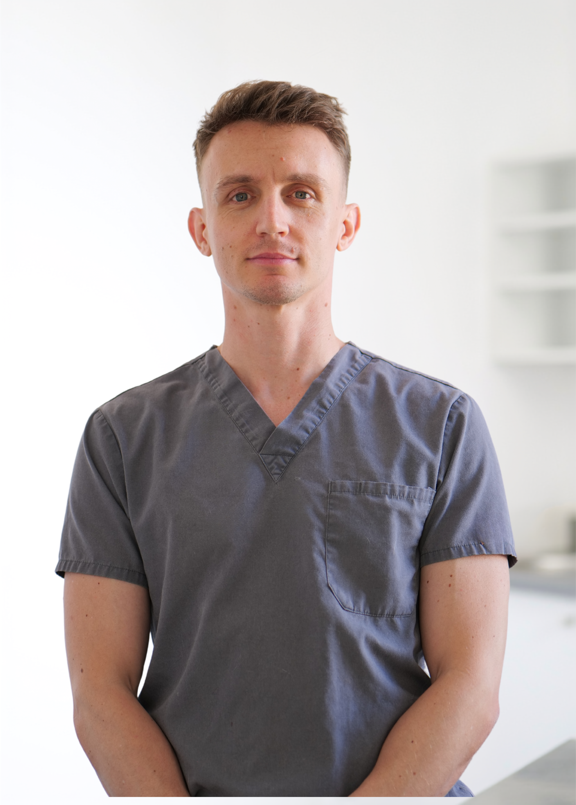 Personne en blouse médicale grise, debout à l'intérieur sur fond clair, face à la caméra.