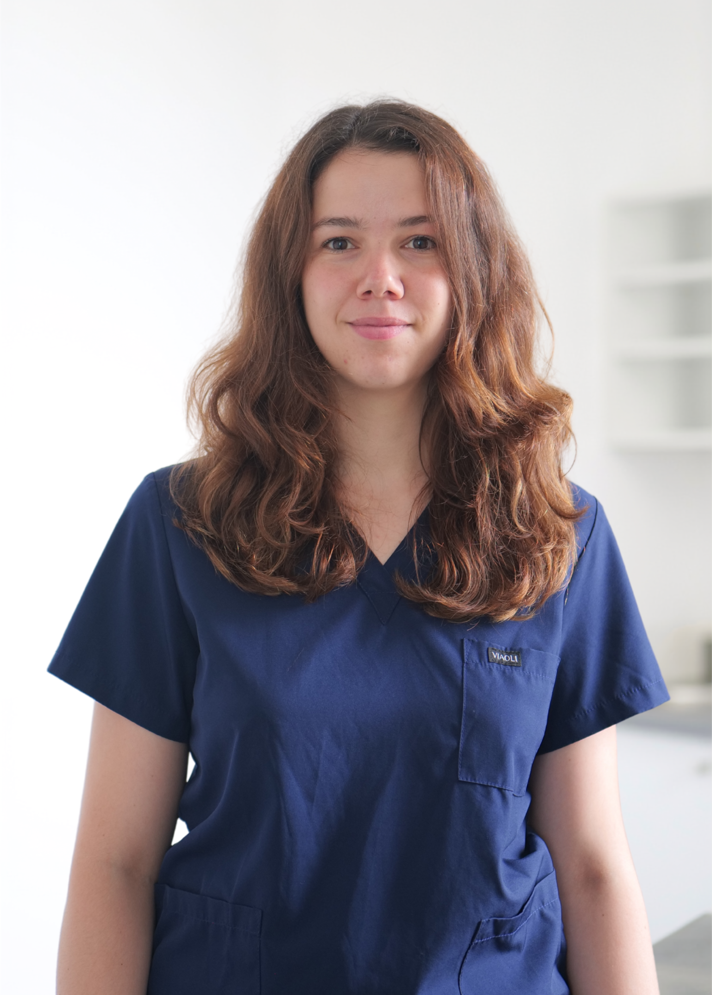 Femme en blouse médicale bleu marine, debout à l'intérieur sur fond clair, regardant droit devant elle avec une expression neutre.