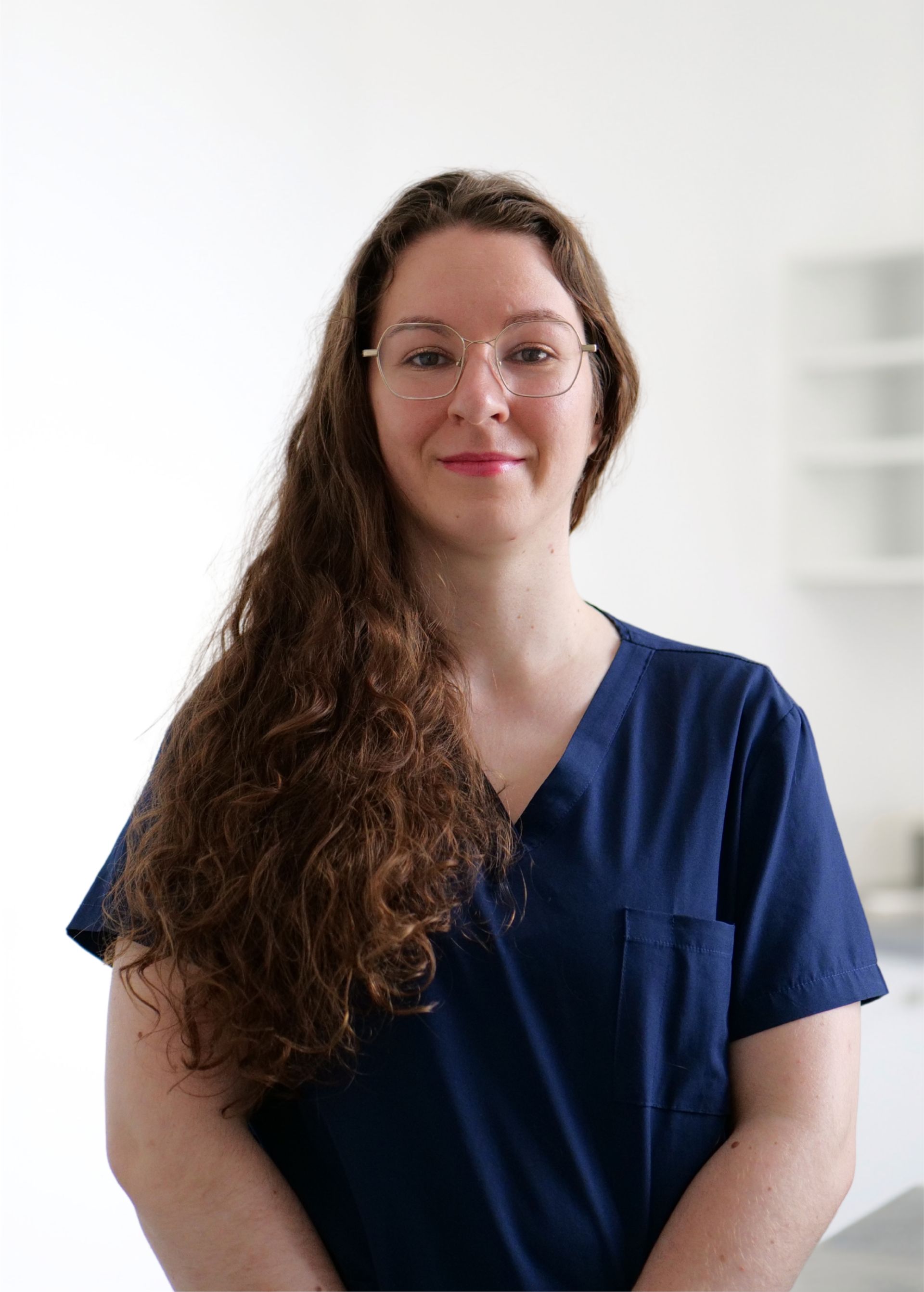 Personne en blouse médicale bleu marine, debout sur fond blanc, esquissant un sourire.