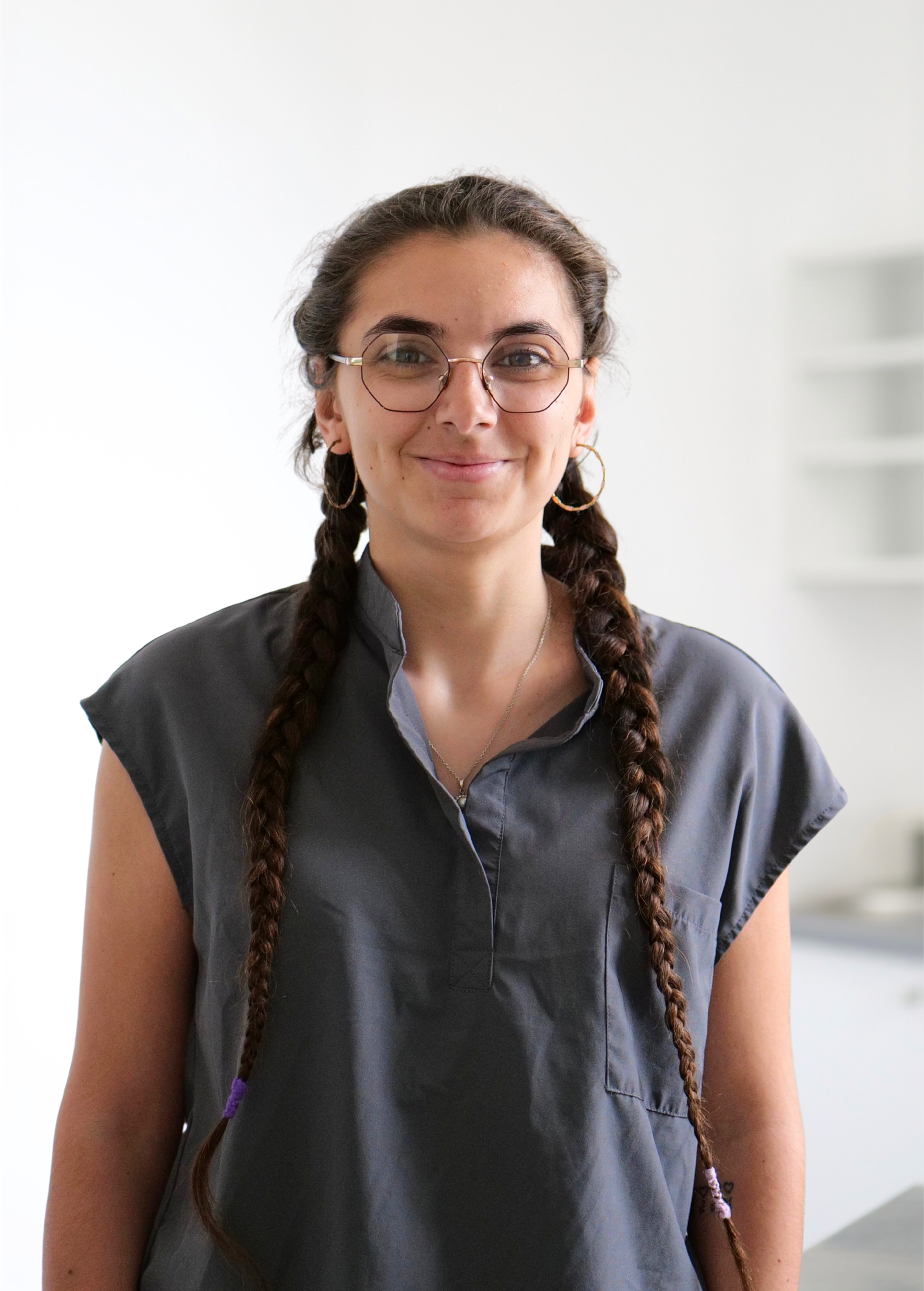 Personne portant des lunettes et des tresses, vêtue d'une chemise foncée, se tenant à l'intérieur sur un fond clair.