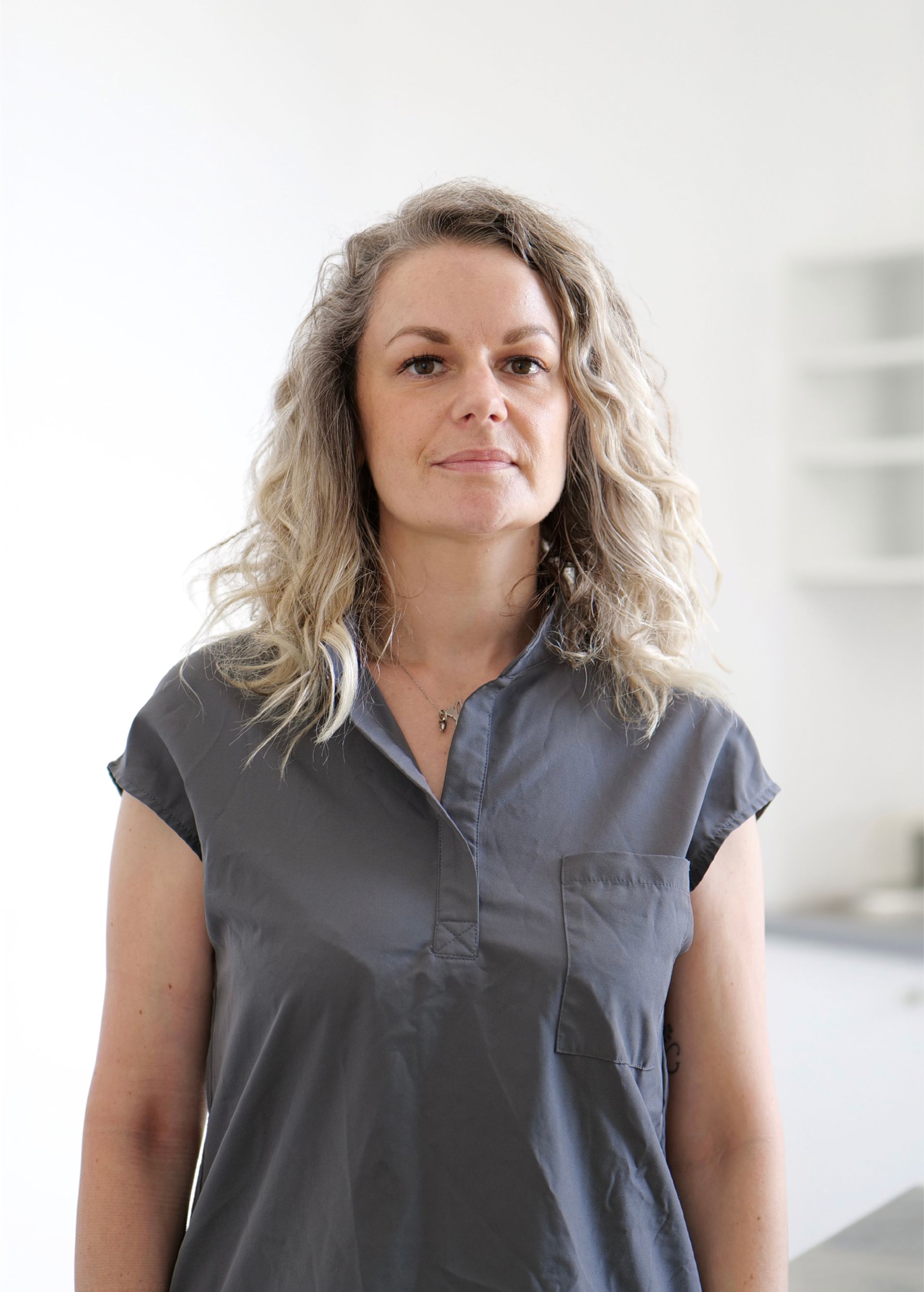 Personne debout à l'intérieur, vêtue d'un chemisier gris, regardant droit devant elle avec une expression neutre.