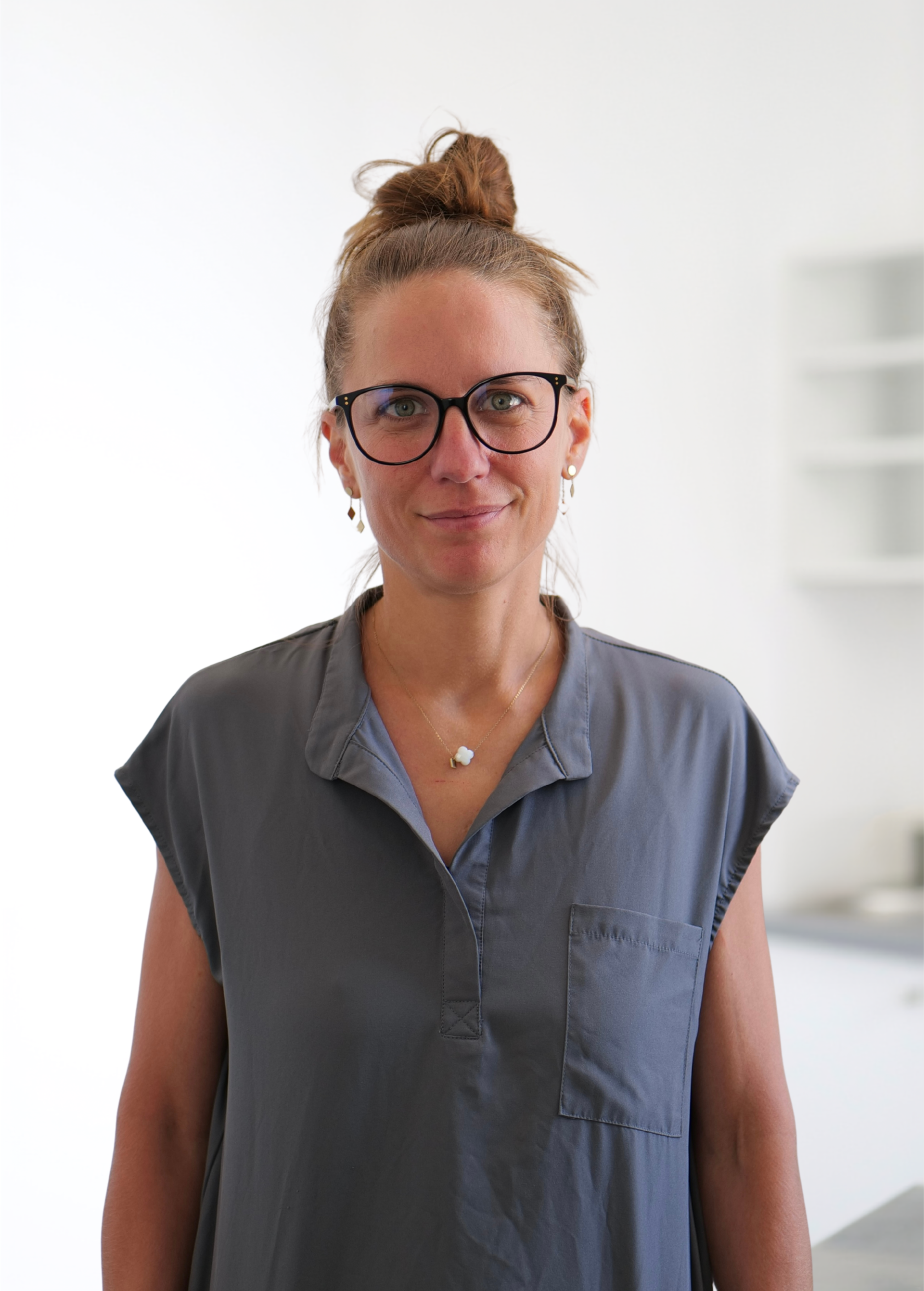 Personne portant des lunettes, les cheveux relevés en chignon, vêtue d'une chemise grise, debout à l'intérieur et faisant face à la caméra.
