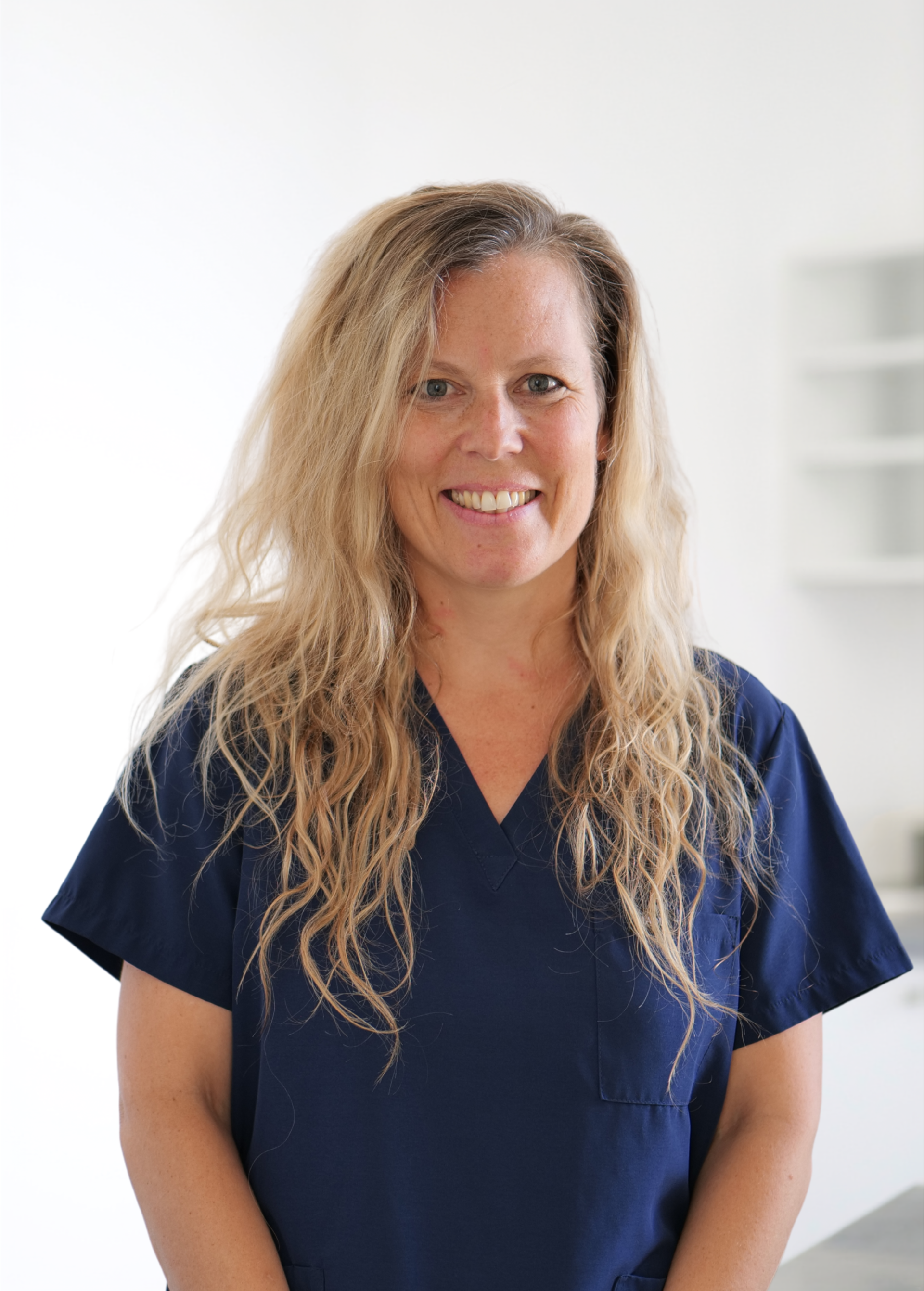 Femme en blouse médicale bleu marine, debout sur fond blanc, souriant à l'objectif.