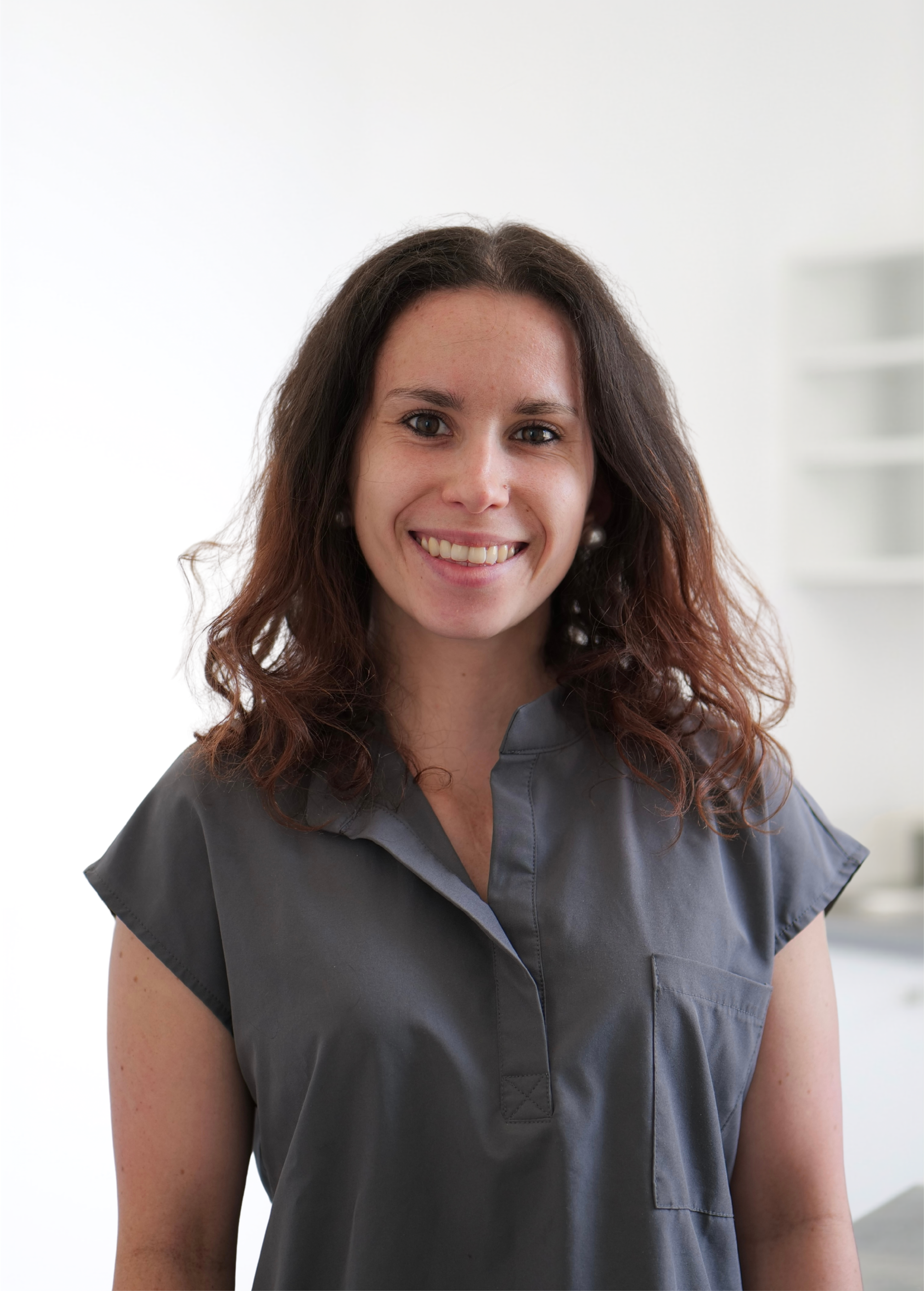 Femme souriante aux cheveux bouclés, vêtue d'un chemisier gris, se détachant sur un fond blanc éclatant.