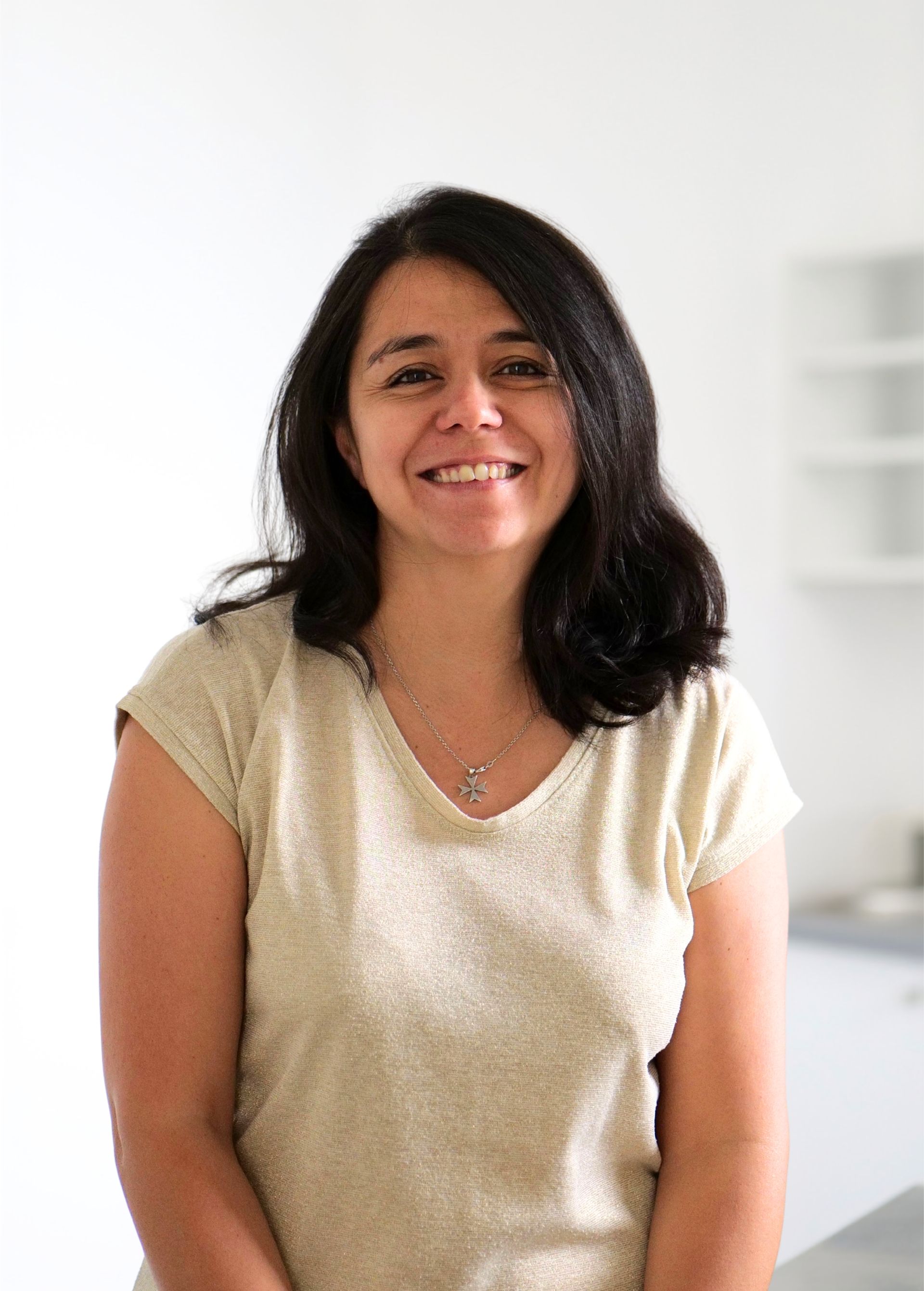 Personne souriante aux cheveux foncés, vêtue d'un haut beige clair, se tenant à l'intérieur sur un fond blanc éclatant.