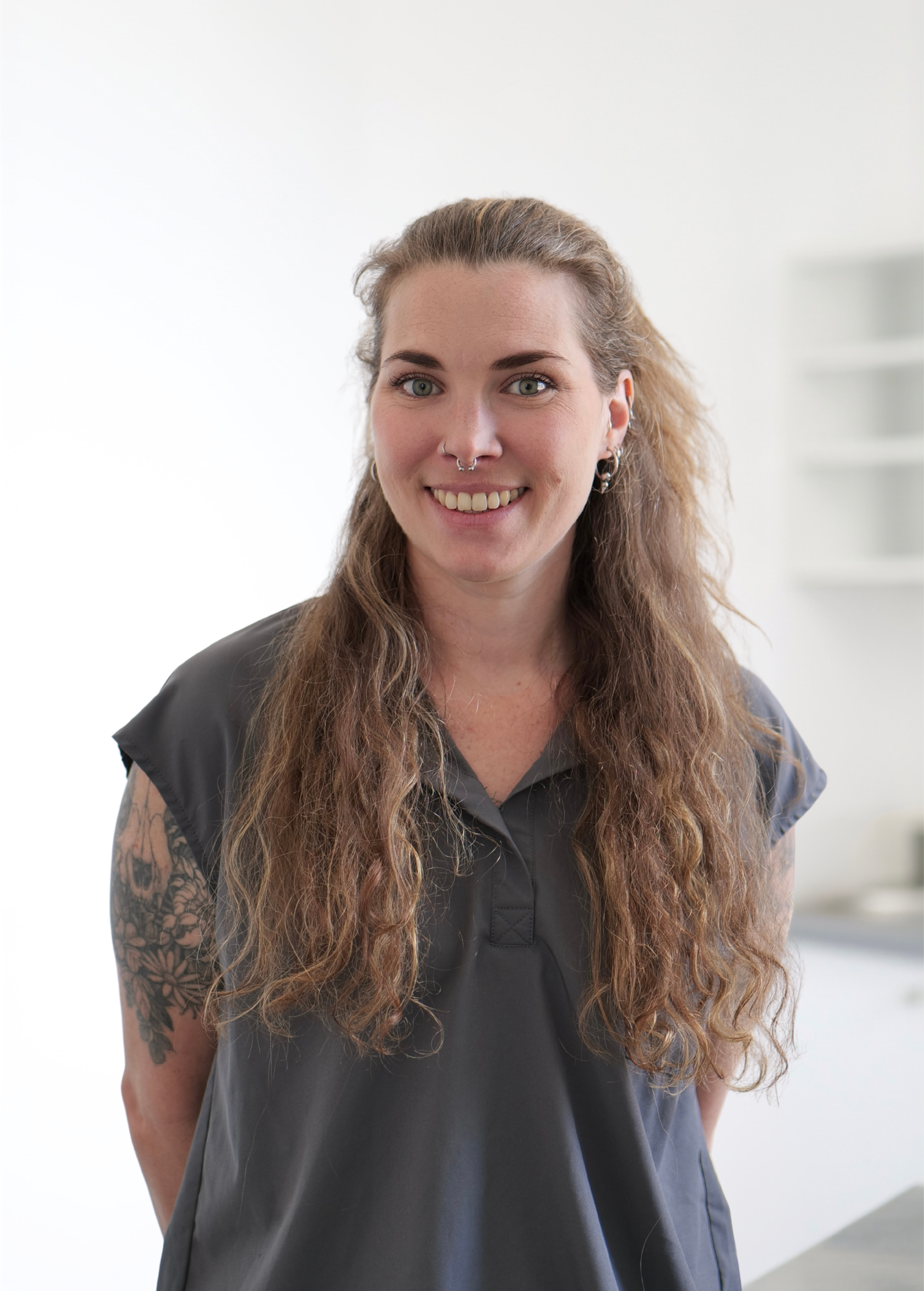 Personne souriante aux longs cheveux bouclés, portant une chemise foncée, se tenant à l'intérieur sur un fond blanc lumineux.