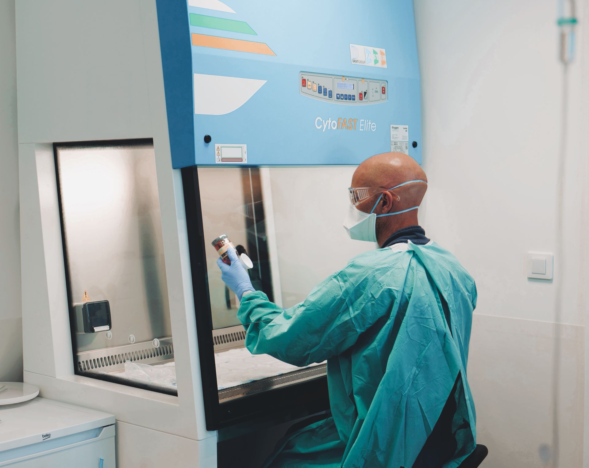 Technicien en combinaison de protection turquoise travaillant à l'intérieur d'une boîte à gants de laboratoire dans une salle blanche.