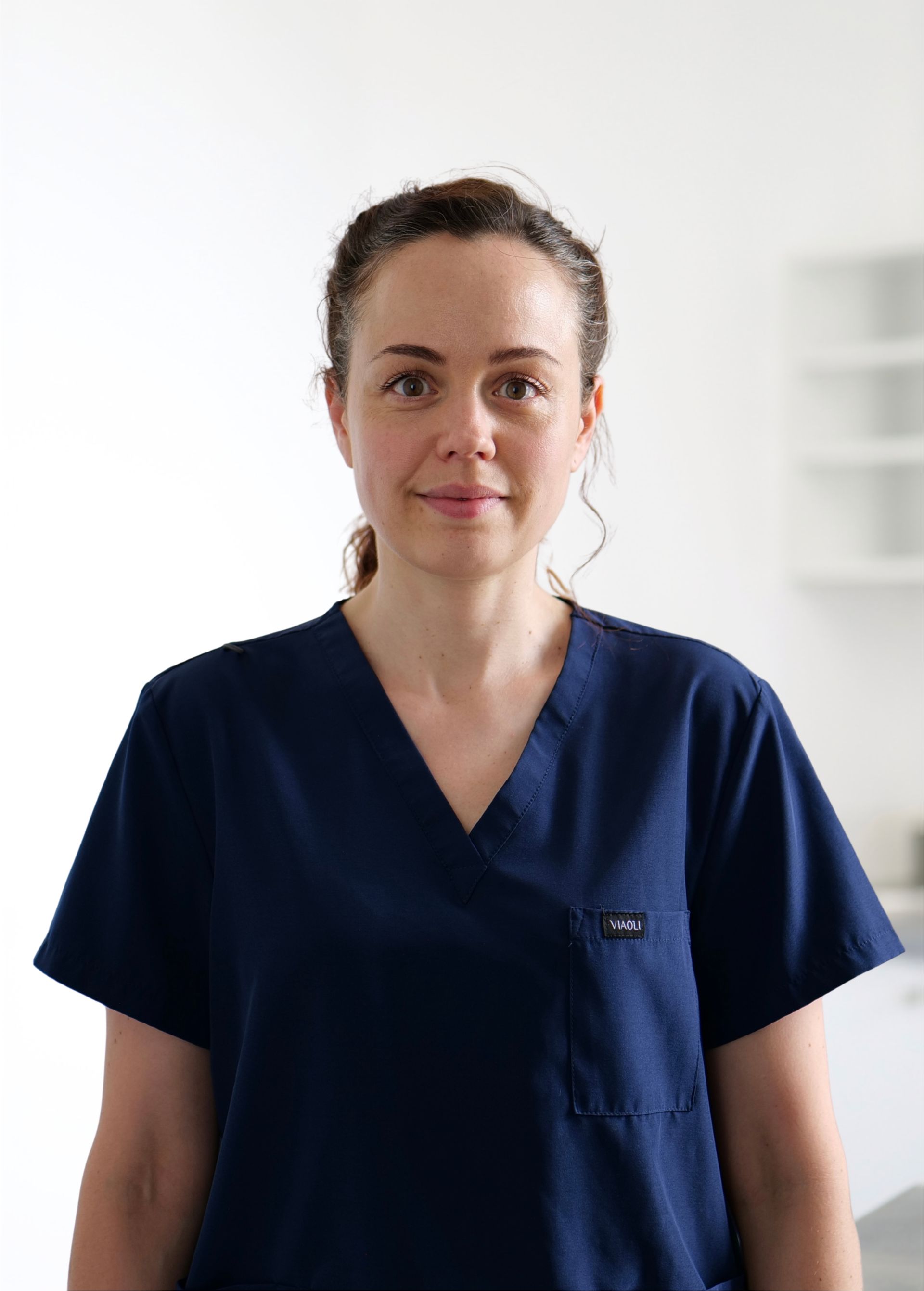 Personne en blouse médicale bleu marine, debout sur fond blanc, regardant droit devant elle avec un léger sourire.