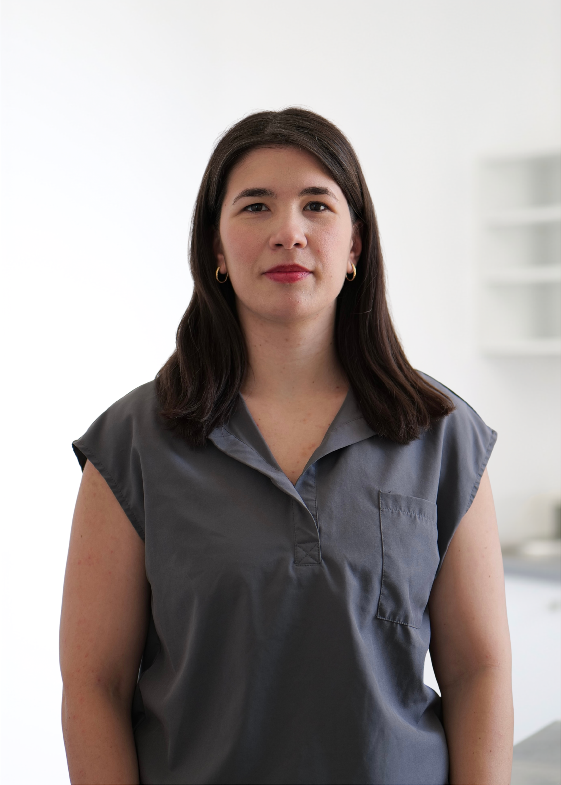 Femme debout à l'intérieur, sur fond blanc, portant un chemisier gris et regardant l'objectif.