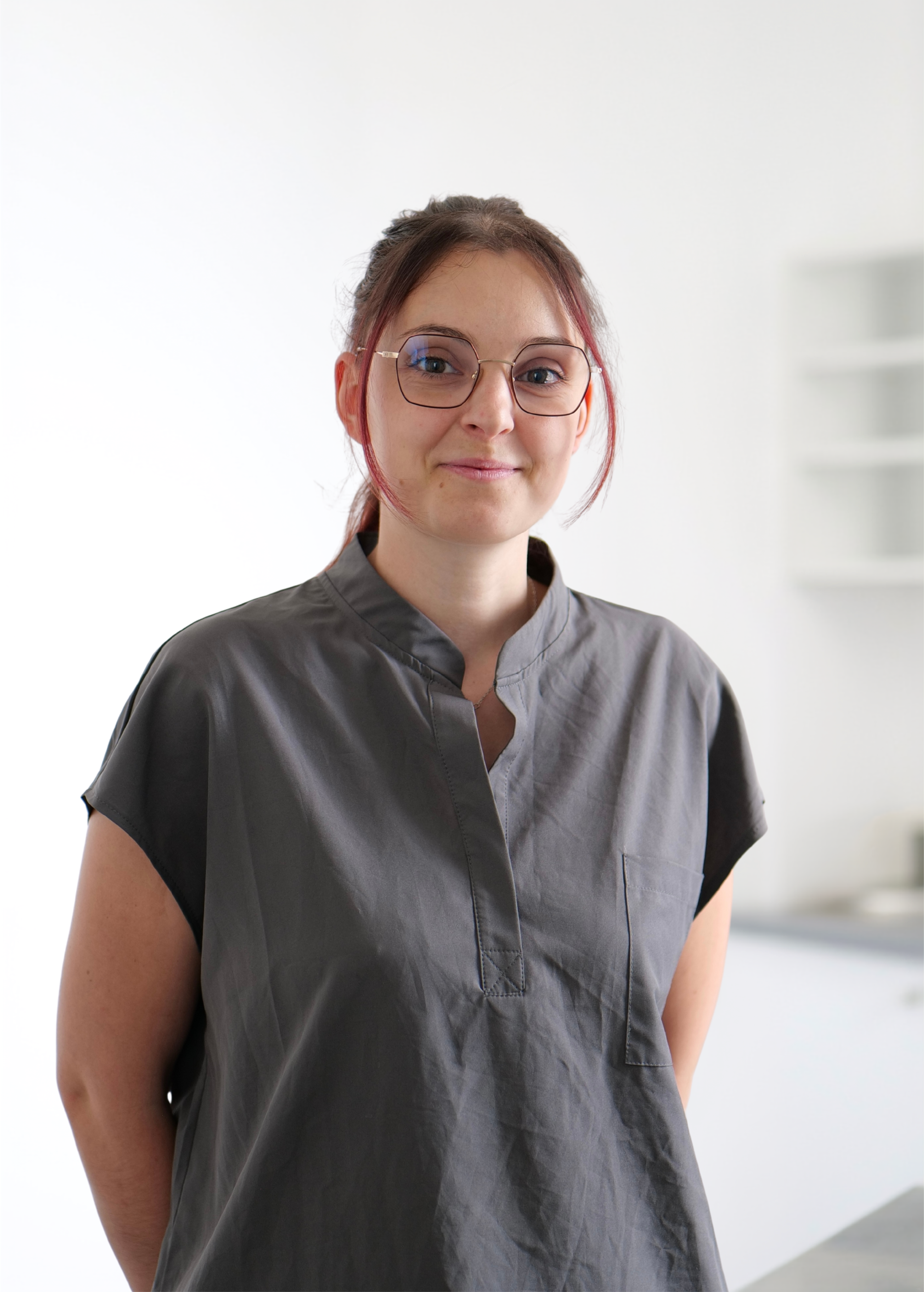 Une personne portant des lunettes et une chemise gris foncé se tient à l'intérieur, sur un fond blanc éclatant.