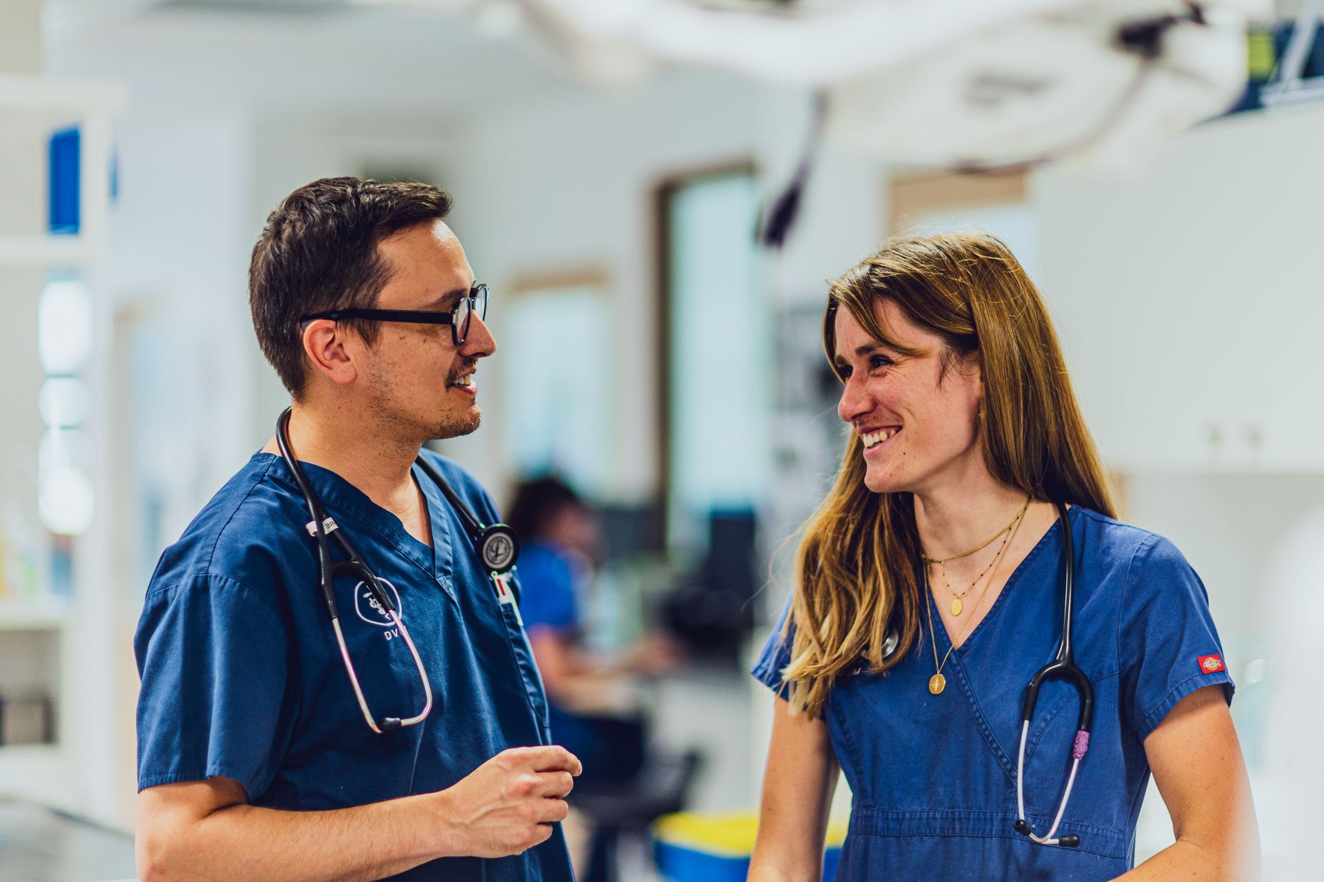Deux membres du personnel médical en blouse bleue discutent dans un couloir d'hôpital, se souriant l'un à l'autre.