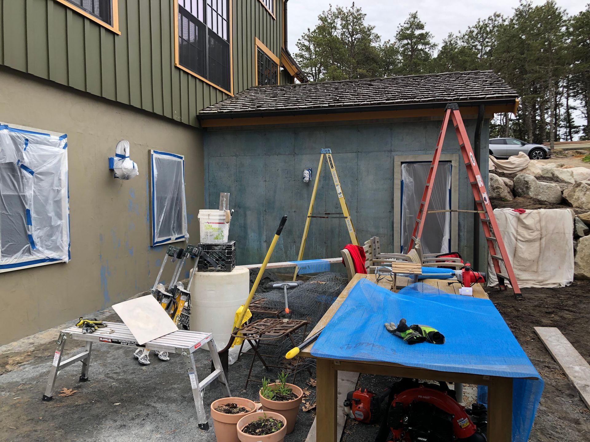 Backyard construction area with ladders, tools, tarp-covered table, and a partially painted house wall