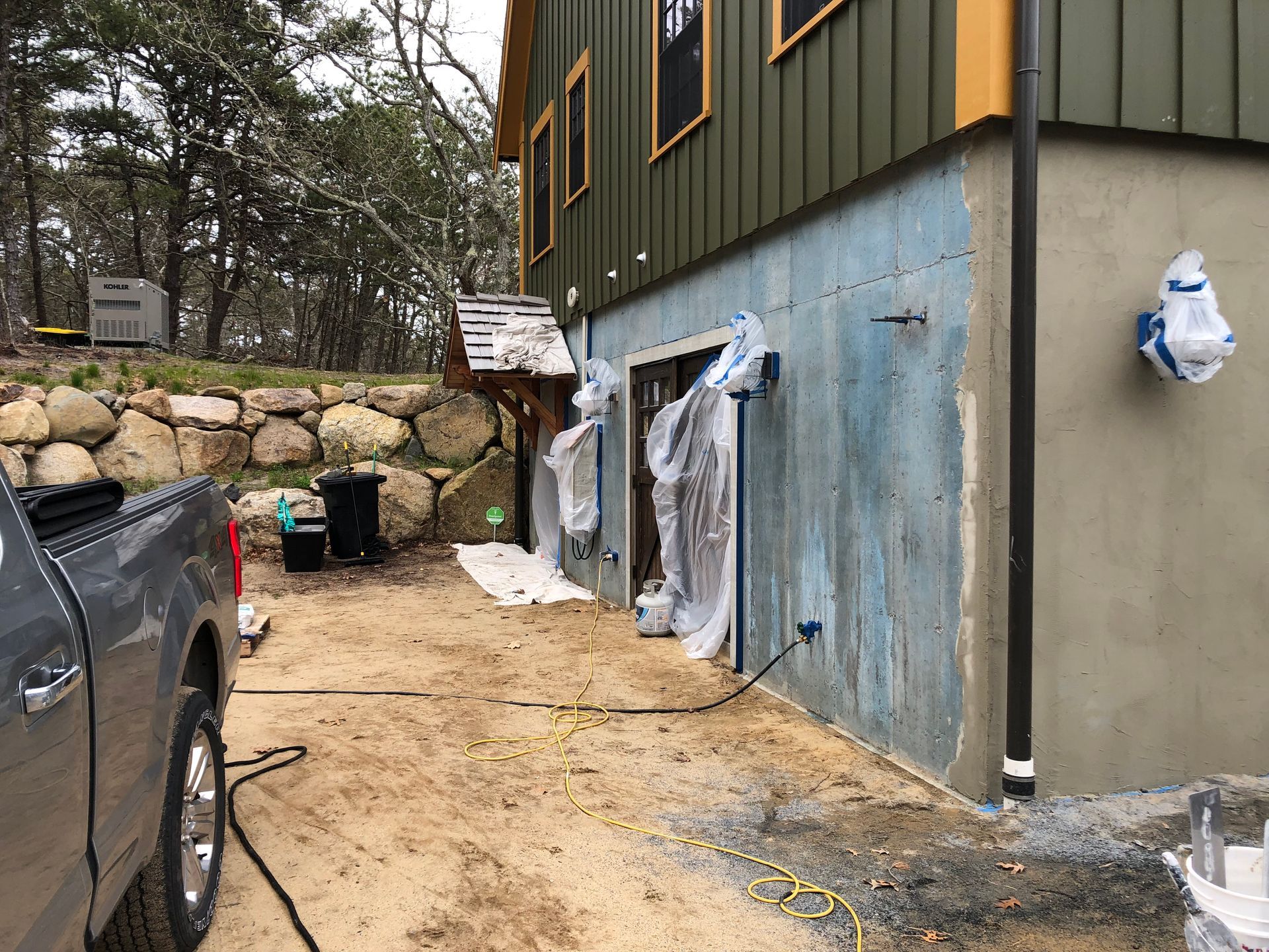 Exterior of a house under construction with covered windows, stone wall, truck, and muddy driveway