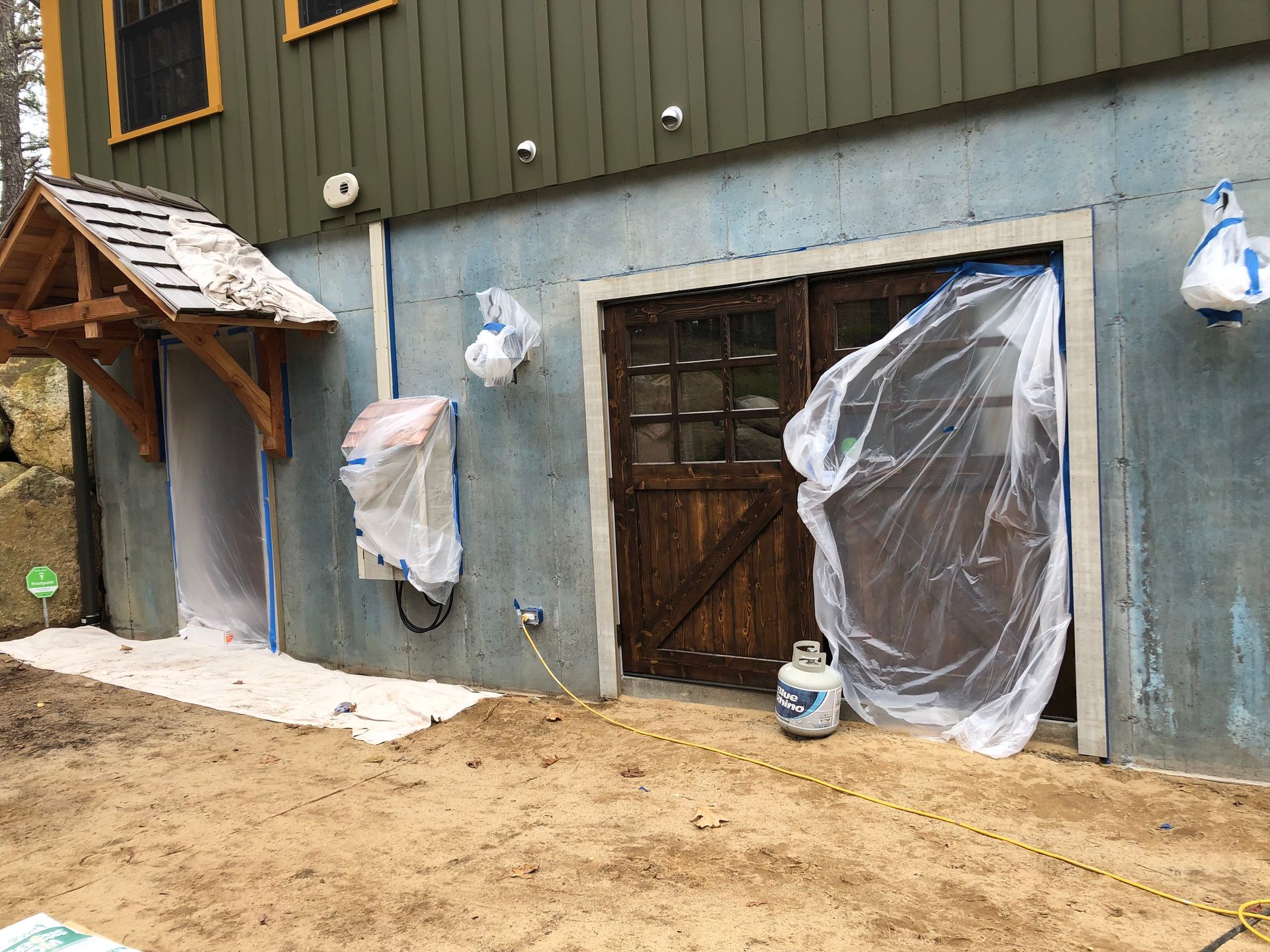 Partially renovated house exterior with plastic-covered openings and sandy ground in front