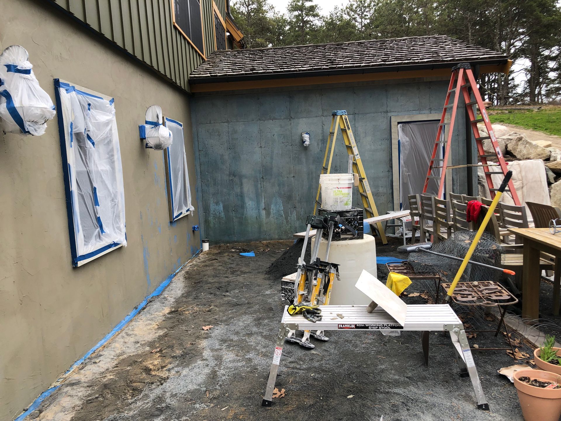 Construction tools and ladders in a narrow outdoor work area beside a house and garage