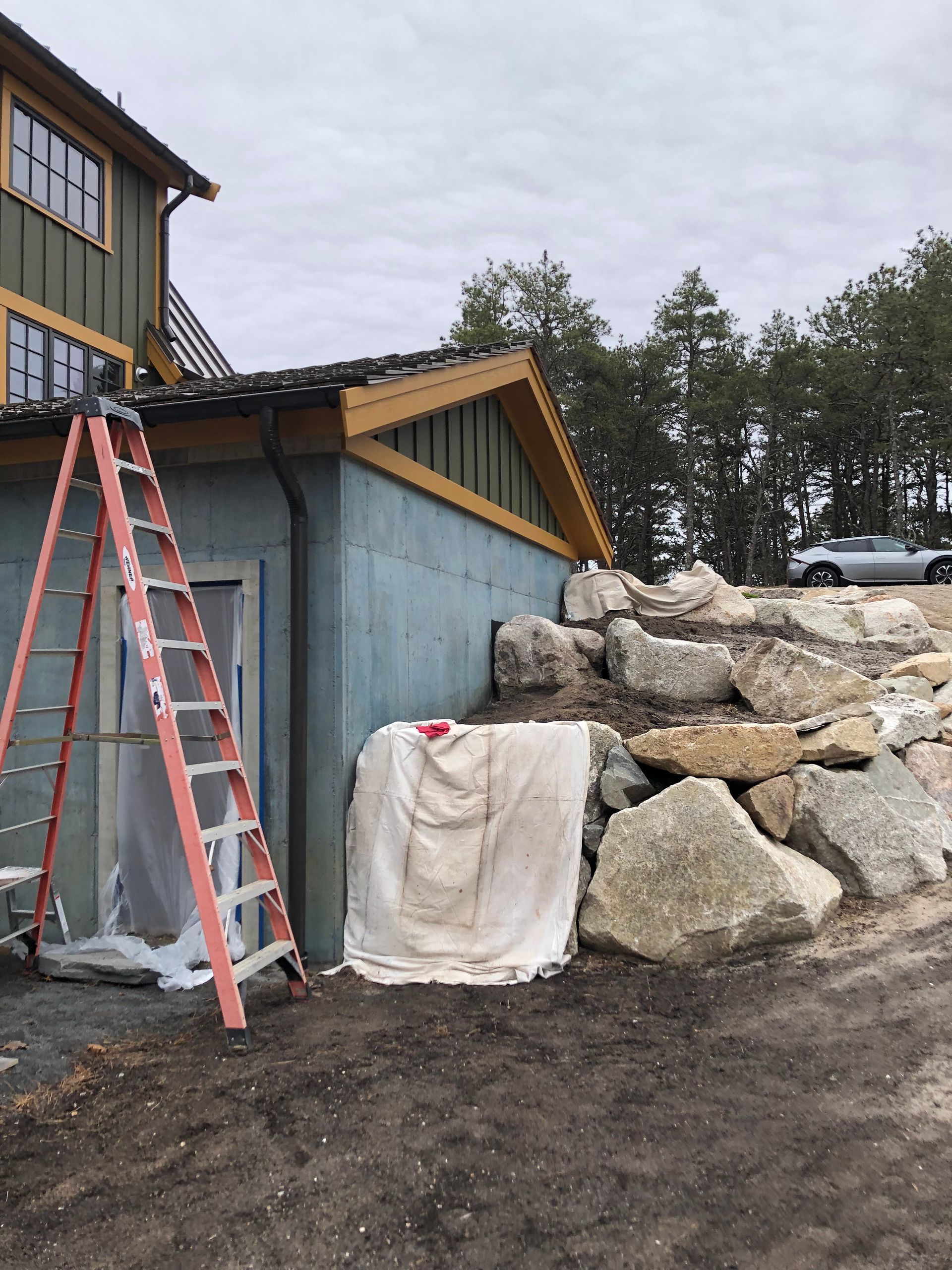 Red ladder beside a blue building and rock retaining wall outdoors under overcast sky