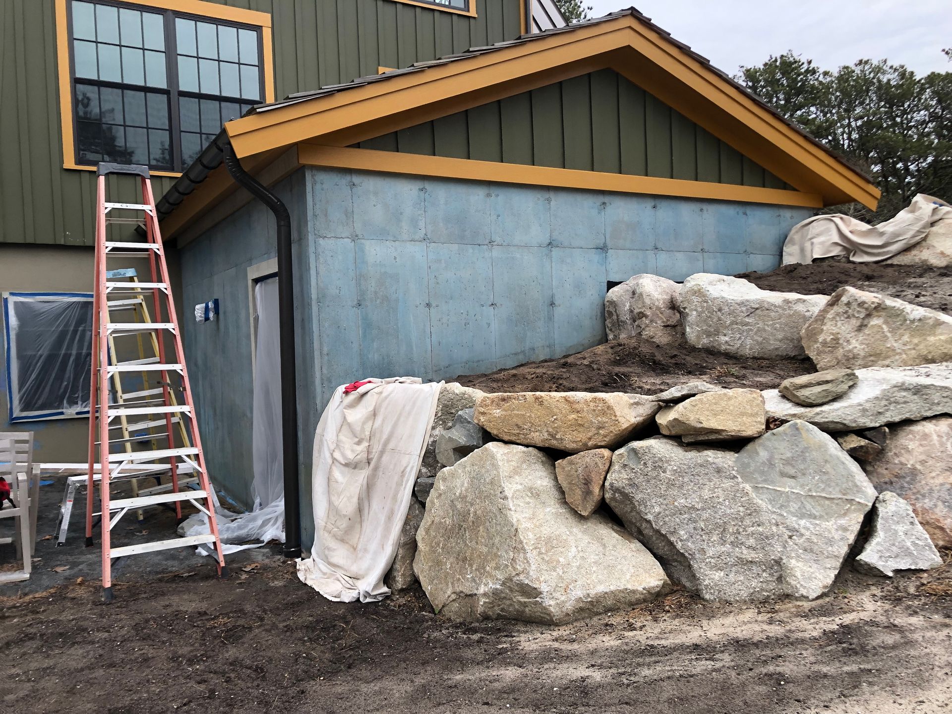 Construction site with blue building wall, ladder, and stacked boulders beside a house