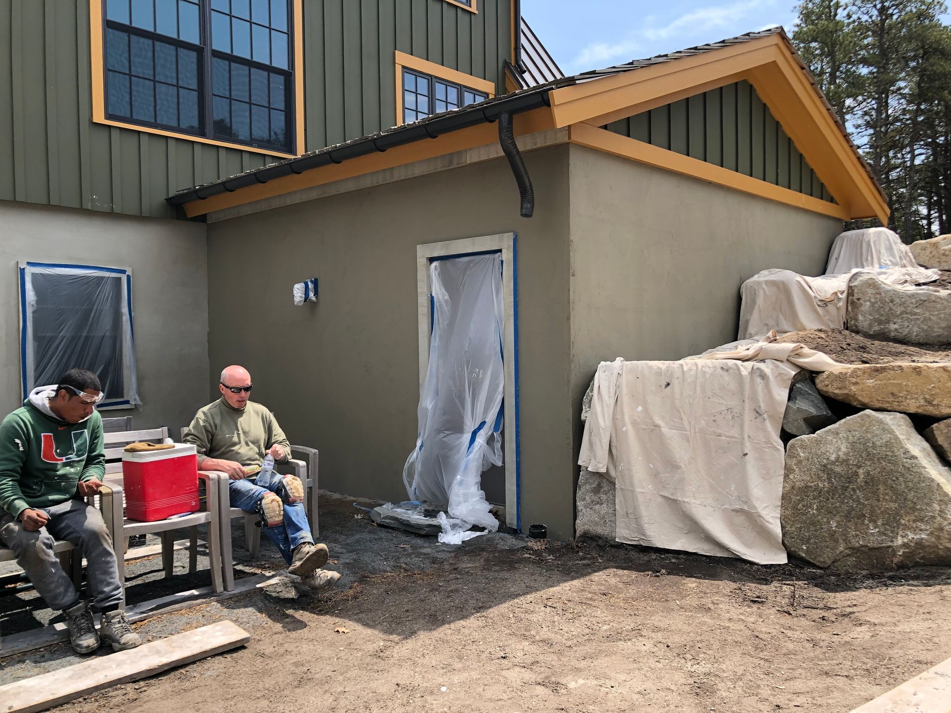 Two workers sitting beside a tan shed under construction, with tarps and stacked rocks nearby