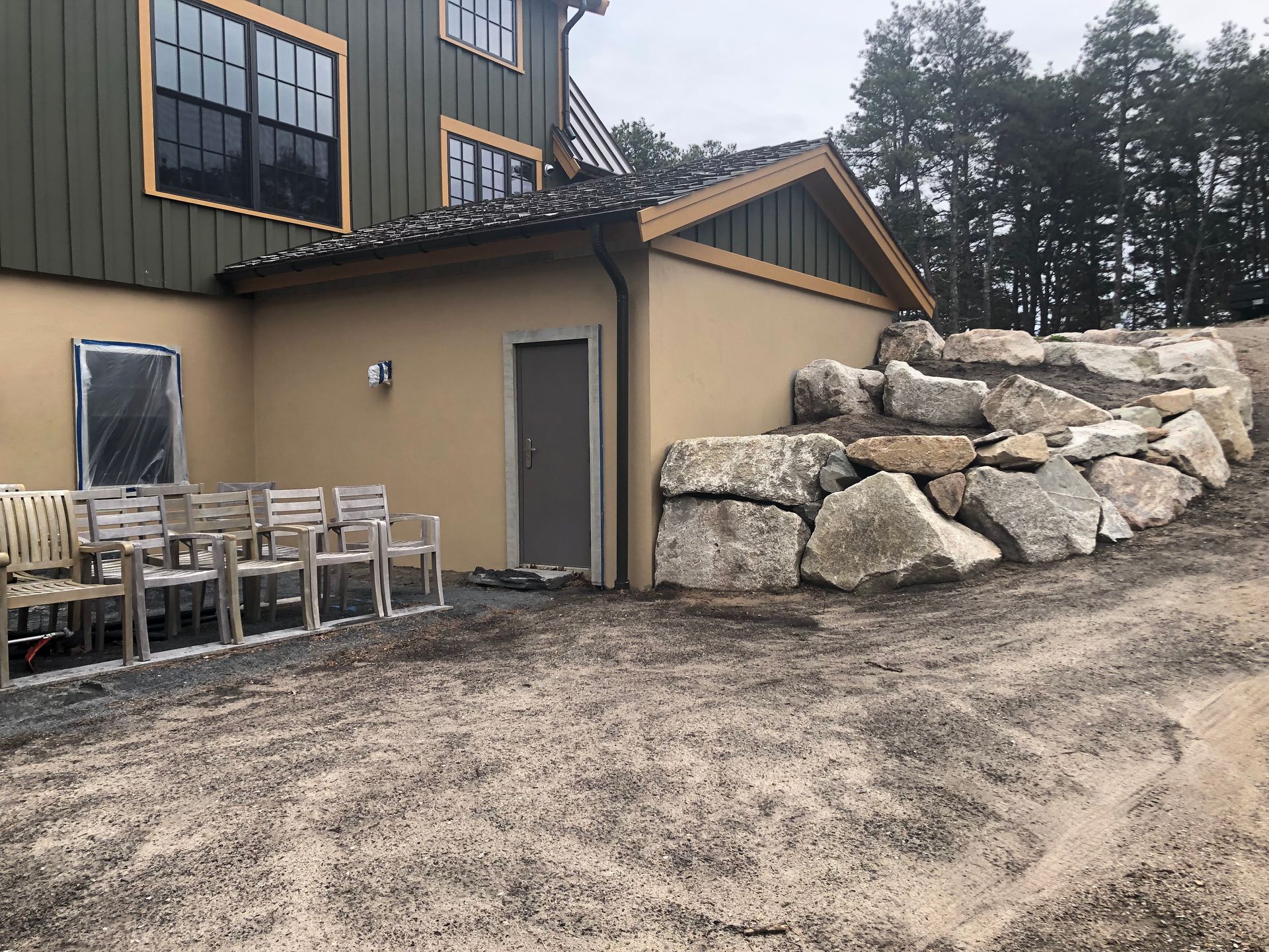 Outdoor patio beside a tan building with stacked chairs and a stone retaining wall under trees
