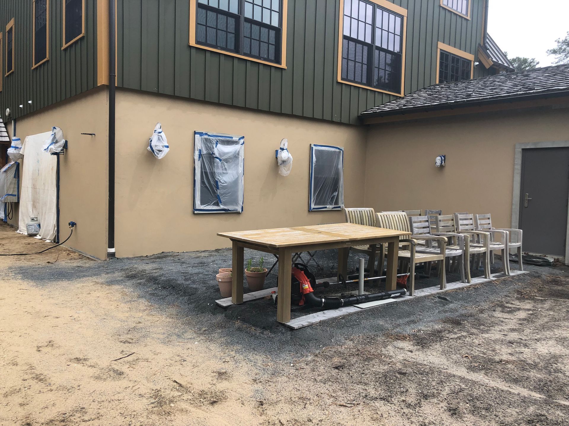 Outdoor patio beside a tan building with a wooden table and stacked chairs on a gravel surface.