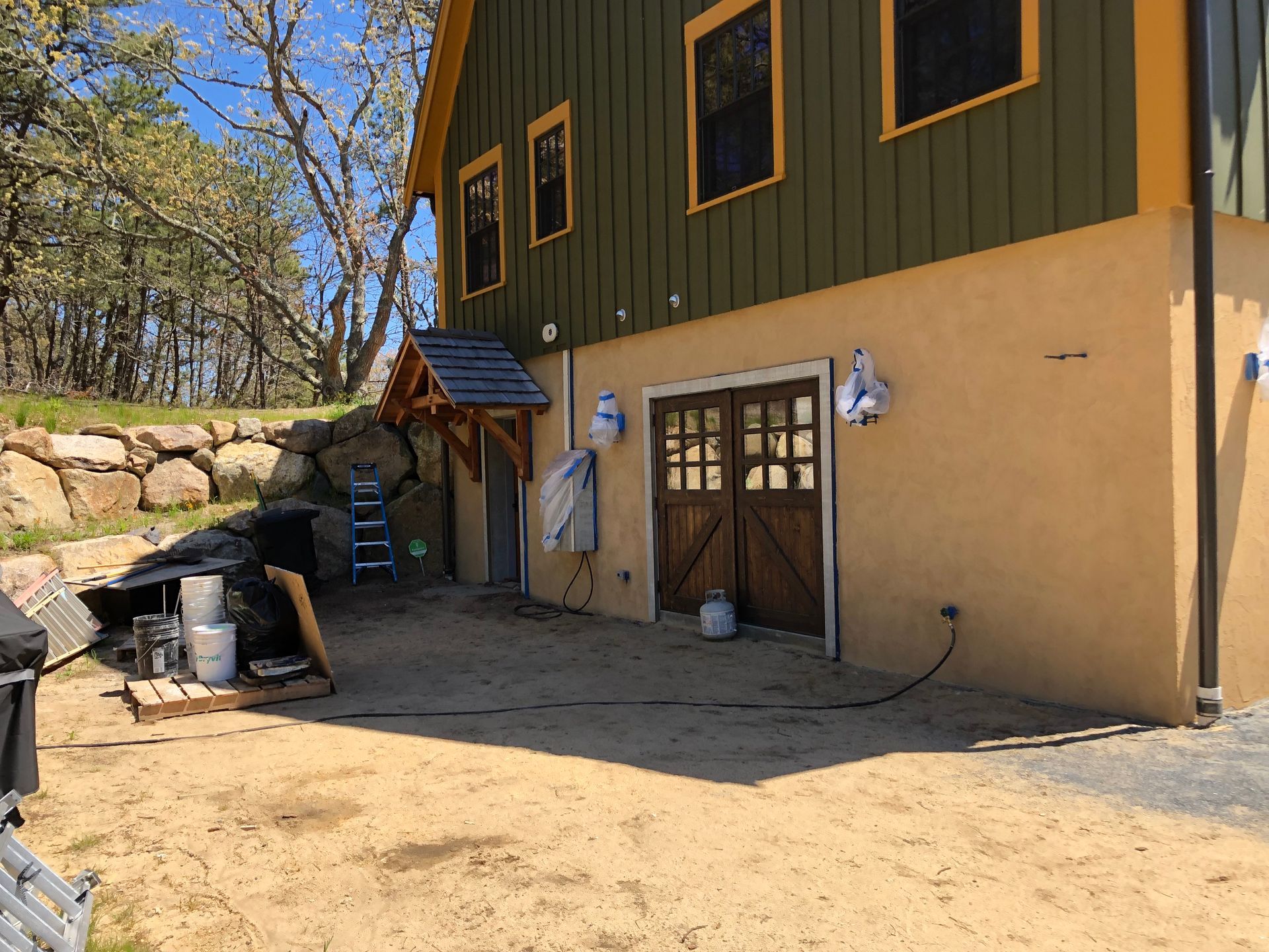 Exterior of a tan and green house with a small wooden shed and yard equipment on a gravel patio
