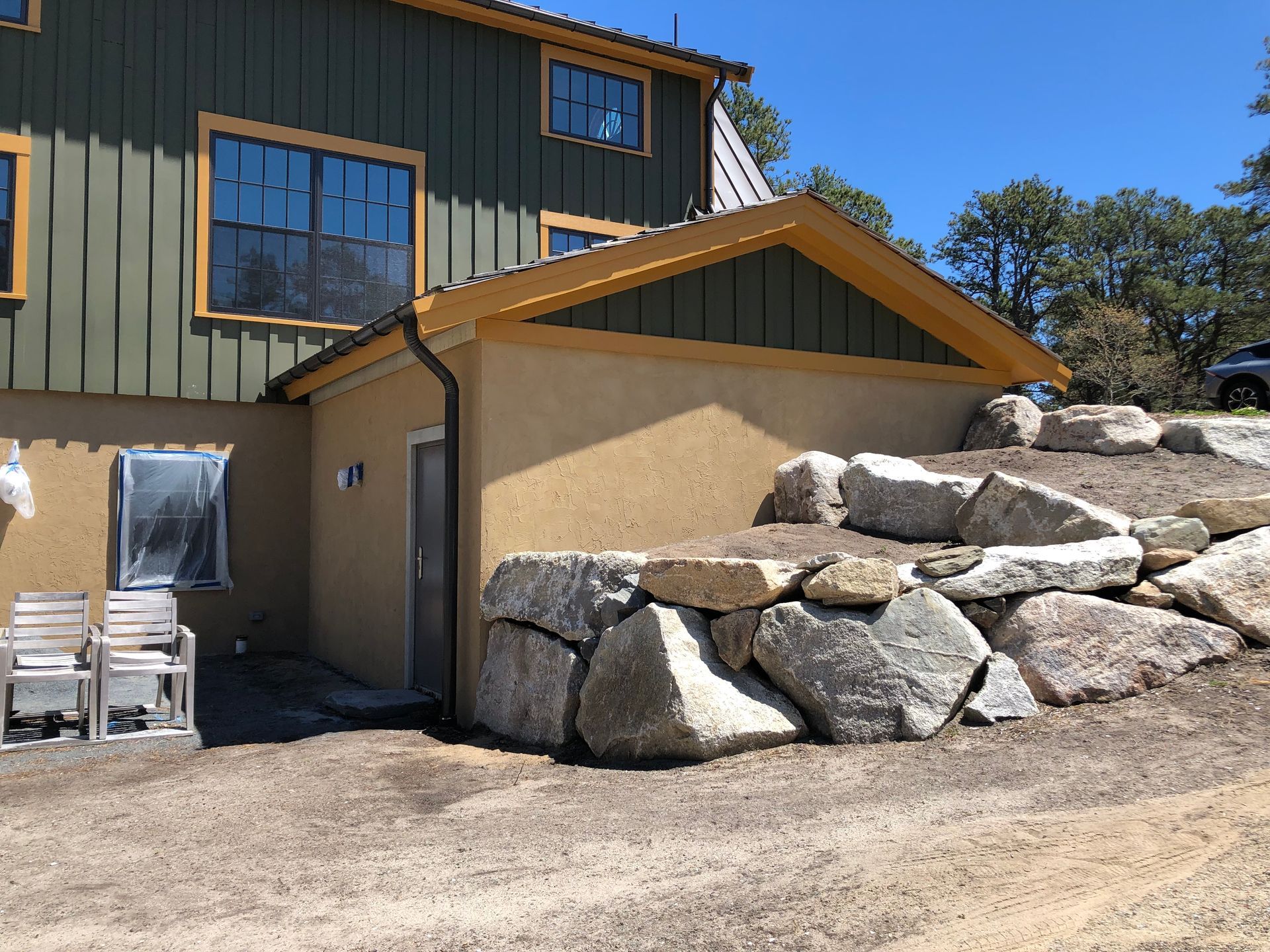 Tan house with large rock retaining wall and white patio chairs under a blue sky